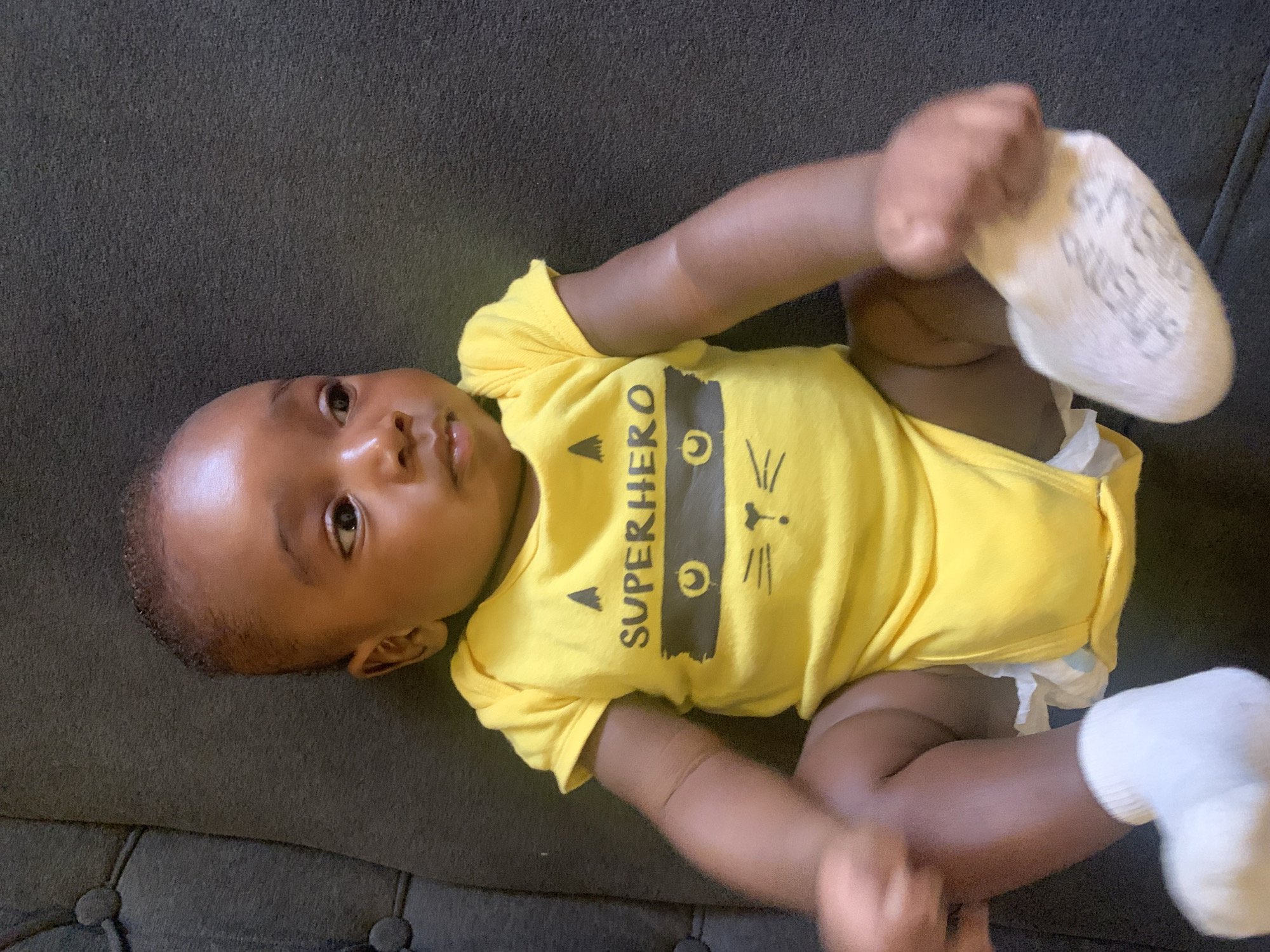 A baby lies on a grey surface. The baby is wearing a yellow onesie with the word "SUPERHERO" printed on it, along with a cat illustration. The baby has dark skin and is wearing white socks. The baby is holding one sock in its hand. A diaper is visible.
I cannot provide the location of this image in the city of Barakin Akawo, Nigeria because the image does not provide information that allows that level of specificity.