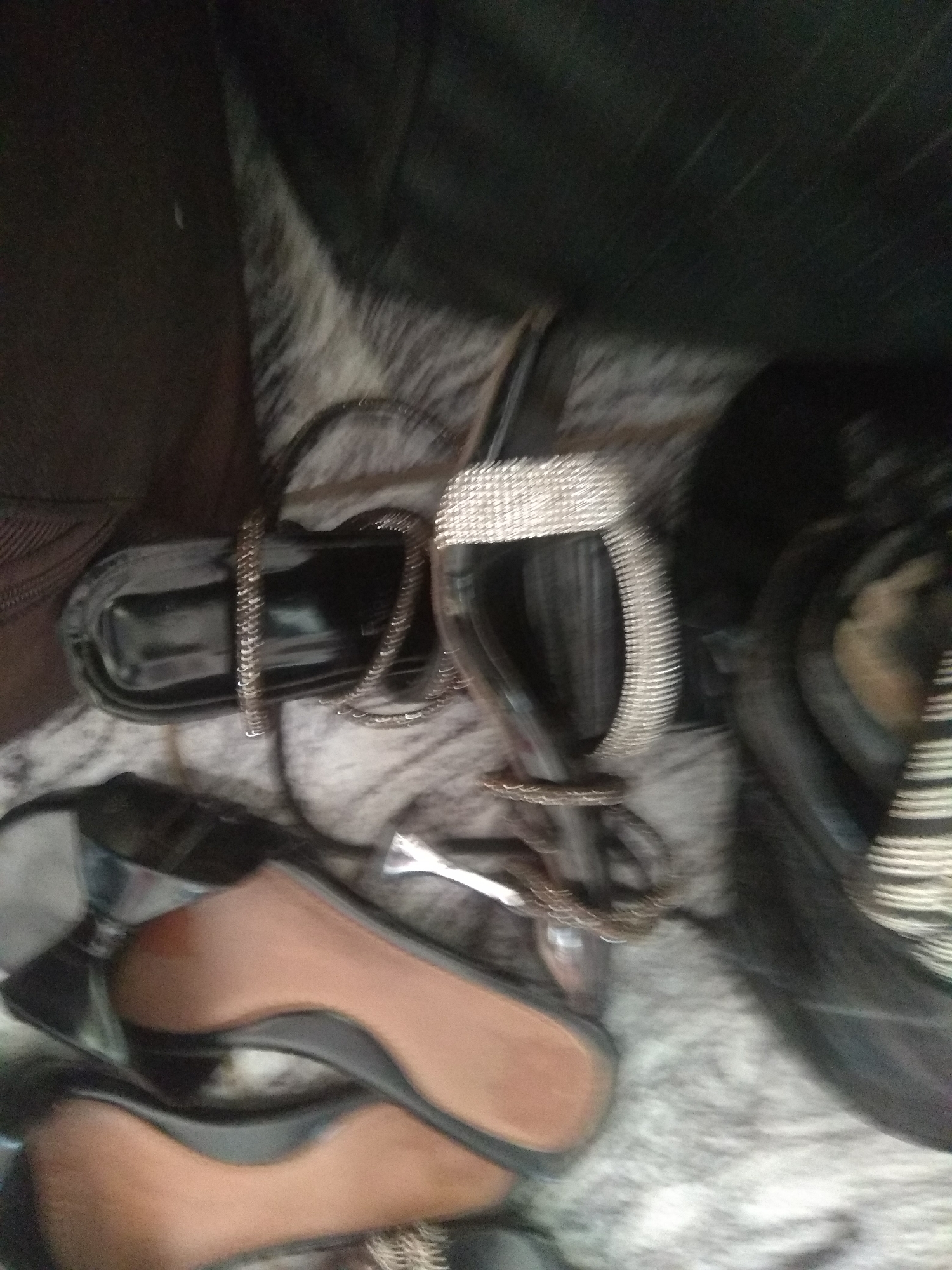 The image displays a collection of women's footwear, including sandals and what appear to be ballet flats, scattered on a textured surface. One pair of black sandals is particularly visible, adorned with a wide, shimmering band across the toe and a delicate, chain-like strap. The soles of some shoes are a tan color, contrasting with the dark uppers. The overall impression is of a casual, indoor setting, possibly a bedroom or closet, where shoes are stored or displayed. The lighting is dim, and the focus is soft, suggesting the photograph was taken indoors with available light. There is no visible text in the image. The context provided indicates the location is Jalingo, Nigeria, but no specific visual cues in the image confirm this.