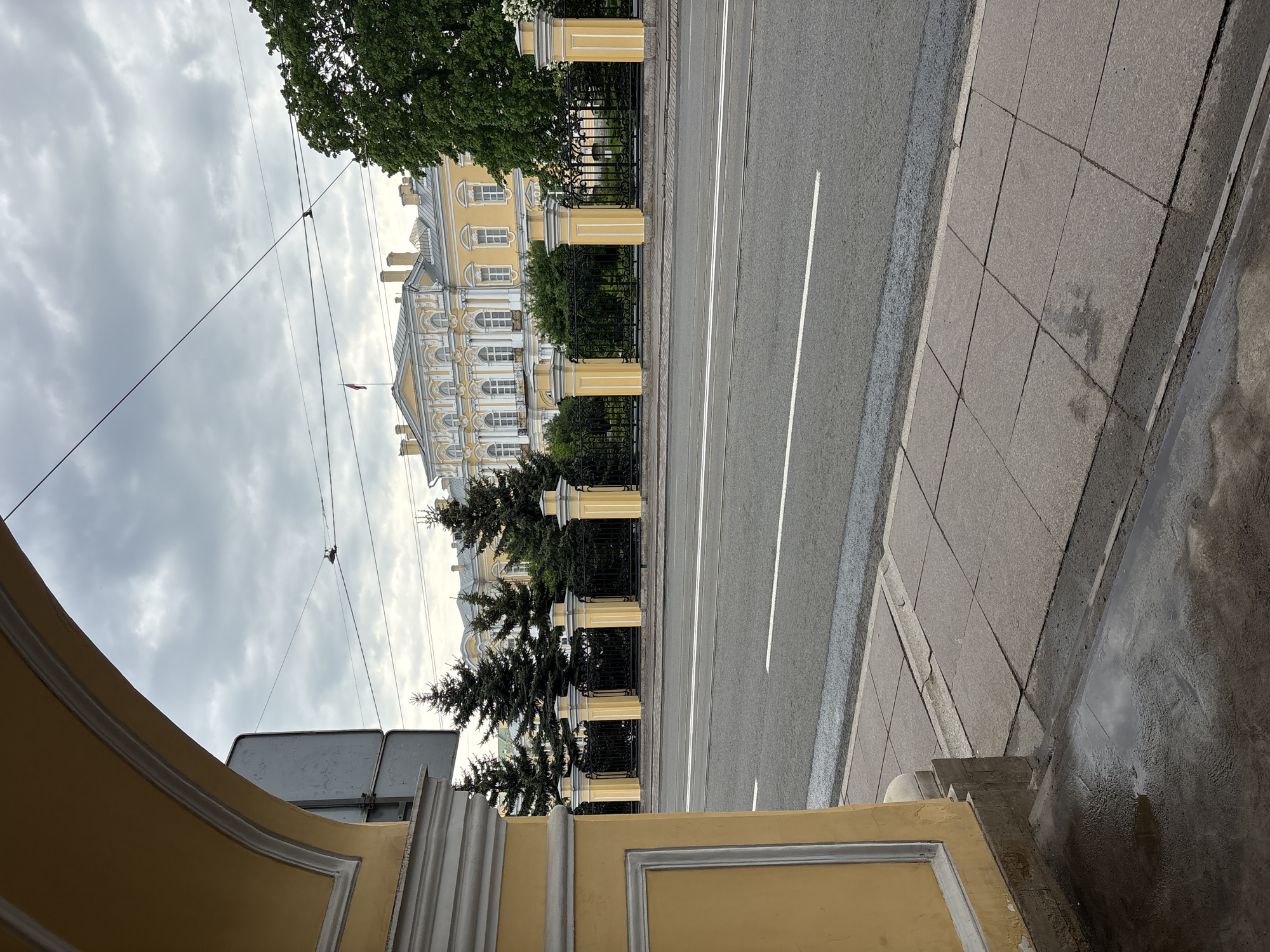 The image depicts a street view in Saint Petersburg, Russia, likely taken near the Smolny Institute. The foreground features a fragment of a yellow building, possibly a wall or part of a façade, with decorative architectural elements. Adjacent to it is a gray tiled sidewalk, with a wet patch suggesting recent rainfall. A gray asphalt road with white lane markings extends into the midground. A decorative black metal fence runs along the road's edge, punctuated by yellow pillars that match the color of the building fragment in the foreground. Behind the fence and adjacent to the road is a row of coniferous trees and green shrubbery. Beyond that, the Smolny Institute is visible, its yellow and white facade a distinct architectural feature. Overhead, thin electrical wires stretch across the sky, and the sky is overcast with gray clouds.