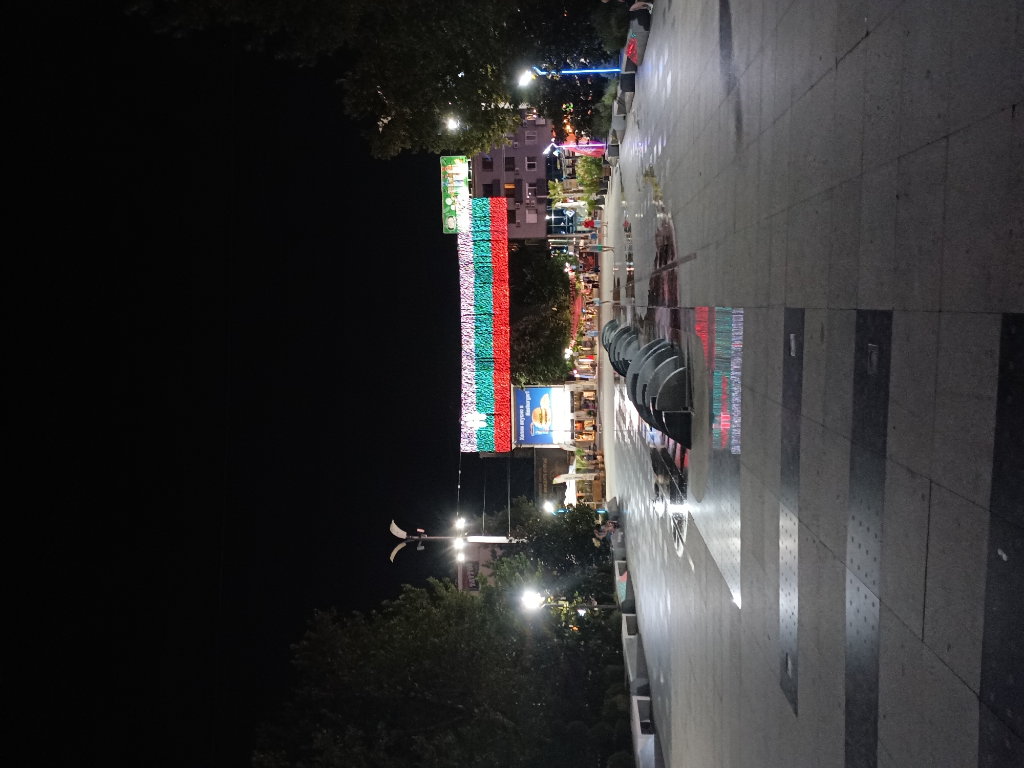 The image depicts a night scene in the city of Burgas, Bulgaria, specifically at the pedestrian bridge connecting the main street to the port. The bridge is illuminated with the Bulgarian flag's colors: white, green, and red. In the foreground, a wet pavement with a fountain feature is visible. Several people are scattered throughout the scene, some sitting on benches, others walking. Buildings and trees line the sides of the plaza. The overall atmosphere is lively and festive, typical of an urban night environment.