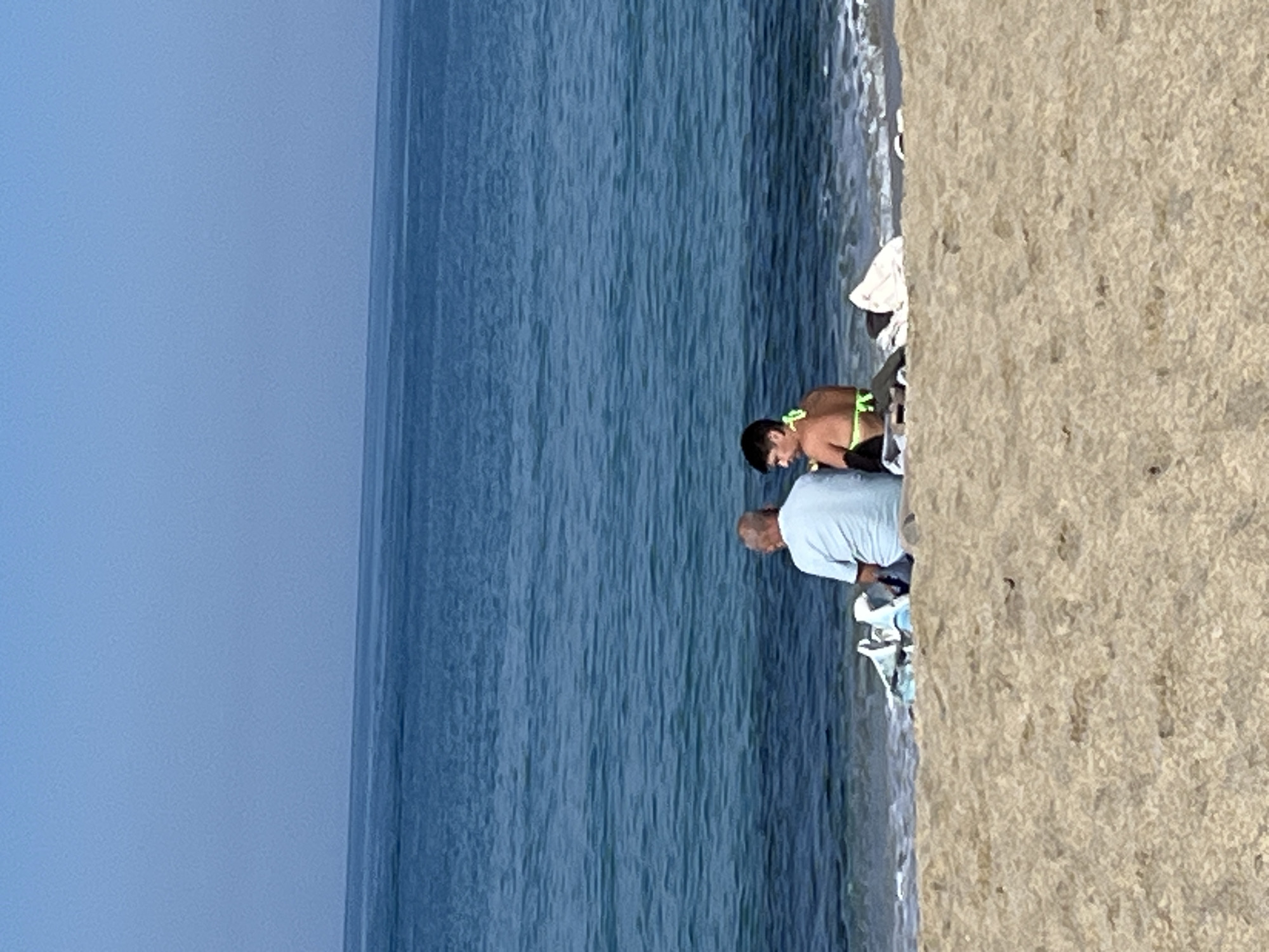 A medium shot depicts a beach in Alanya, Turkey. The image is horizontally oriented. The scene shows a beach with light tan sand to the right, and blue water taking up the center and left of the image. The sky is light blue. Two people are positioned near the shoreline: an older man wearing a light blue shirt and a younger person wearing a black and neon green swimsuit. They are both bending over. There is a small pile of personal belongings including a towel behind them.