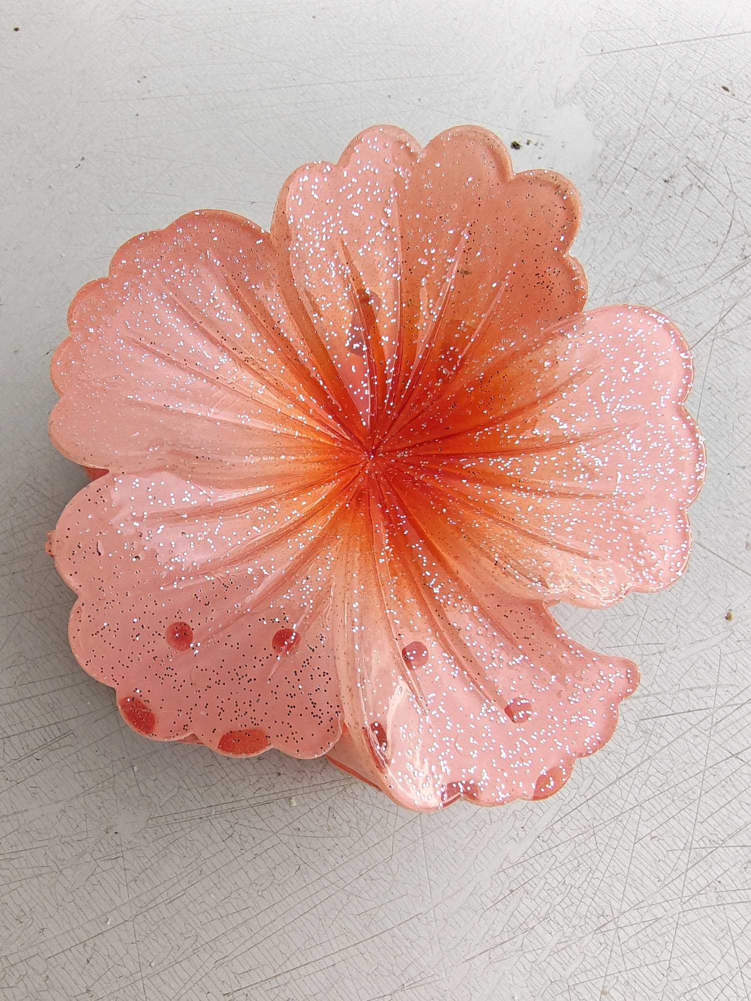A pink and orange glitter-covered hair clip in the shape of a flower rests on a light-colored, textured surface. The flower has rounded, scalloped petals that radiate from a central, orange-hued core. The surface beneath the clip appears to be a table or countertop with a network of fine, light gray lines, possibly cracks or a textured pattern. The image does not provide information about the city of Petroșani, Romania.