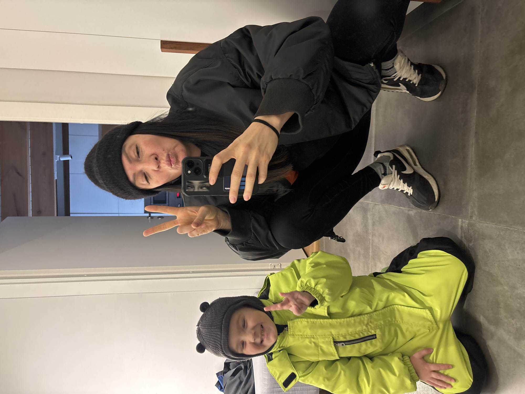 The scene is an interior space within the city of Chaikovsky, Russia. Two individuals, an adult female and a young child, are captured in a mirror selfie. The adult female, positioned on the right, is squatting slightly, wearing a black knit beanie, a black bomber jacket, black leggings, grey socks, and black and white athletic shoes. She holds a dark-cased smartphone in her left hand, angled to capture the reflection, and forms a peace sign with her right hand while puckering her lips. The young child, appearing male and positioned on the left, is kneeling on a dark grey tiled floor. He wears a dark grey knit beanie with two small ear-like protrusions, and a bright neon yellow full-body snowsuit with black knee and lower leg sections. The child smiles broadly with closed eyes and forms a peace sign with his right hand. The immediate background features a plain white wall, and a glimpse of a wooden beam and blue sky through an opening or window is visible on the upper right.