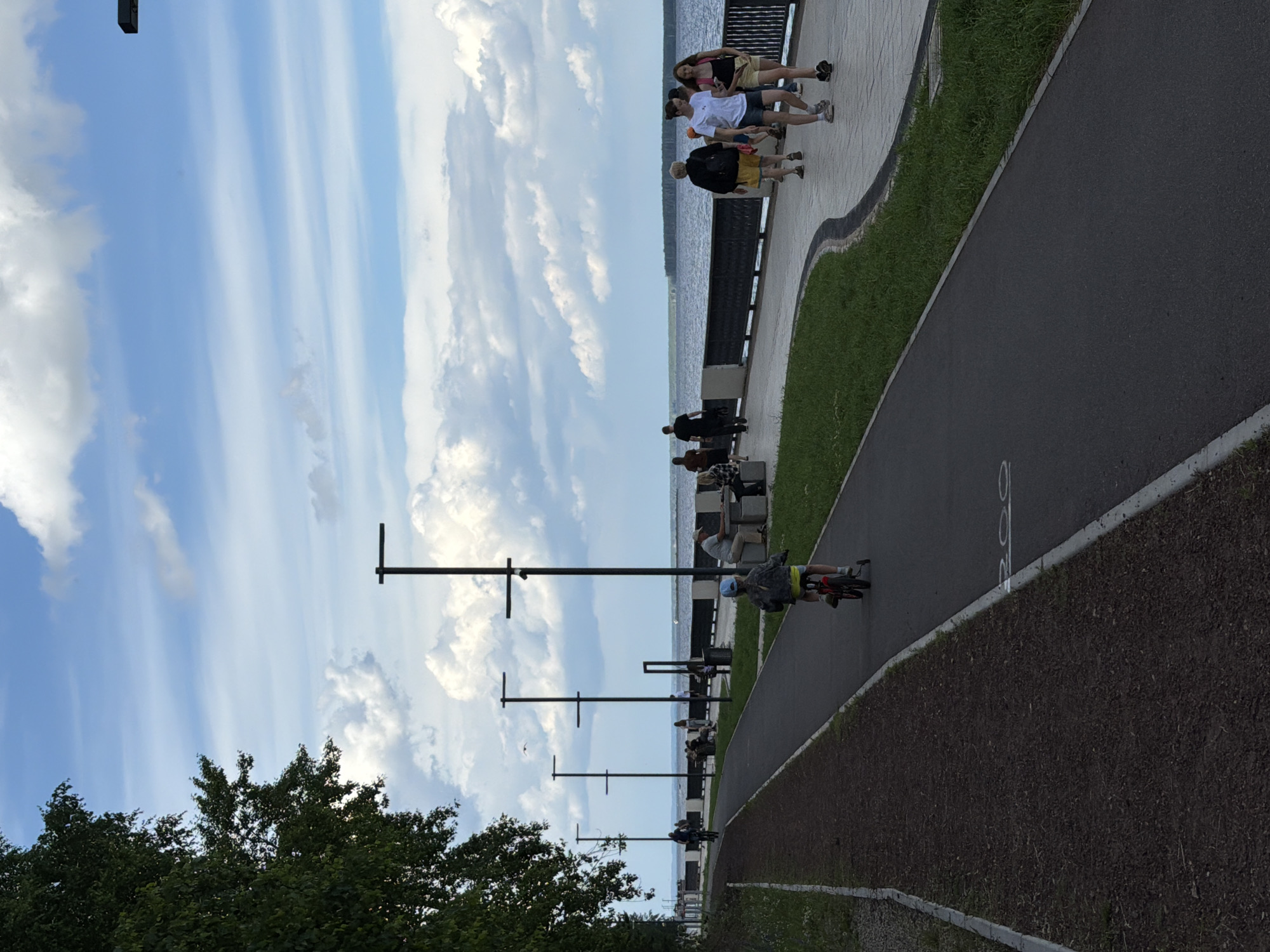 The image depicts a paved walkway and bike path along a waterfront in Chaikovsky, Russia. The sky is partially cloudy with a light blue hue. On the right side of the walkway, several people are walking and interacting. A young child is riding a small bicycle with training wheels on the bike path. Further along the walkway, other individuals are seated on benches. The water body, likely the Kama River, is visible in the background, with a distant tree line on the opposite shore. Modern streetlights are spaced along the walkway, and a dark, treed embankment borders the left side of the scene, separating the paved path from a dirt track. The overall atmosphere suggests a public recreational area on a pleasant day.
