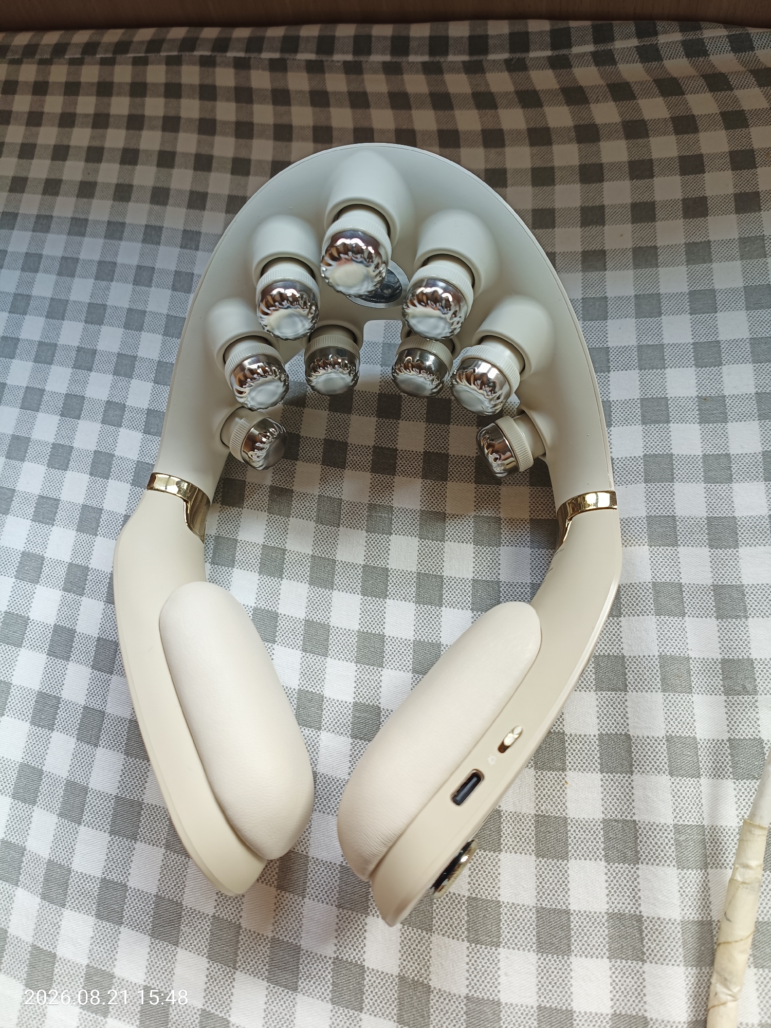 A neck massager with multiple rotating chrome rollers is placed on a grey and white checkered tablecloth. The massager is cream-colored with gold accents. The device has two padded earpieces. A timestamp overlay reads "2026.08.21 15:48". There is no indication of Hong Kong, China in the image.