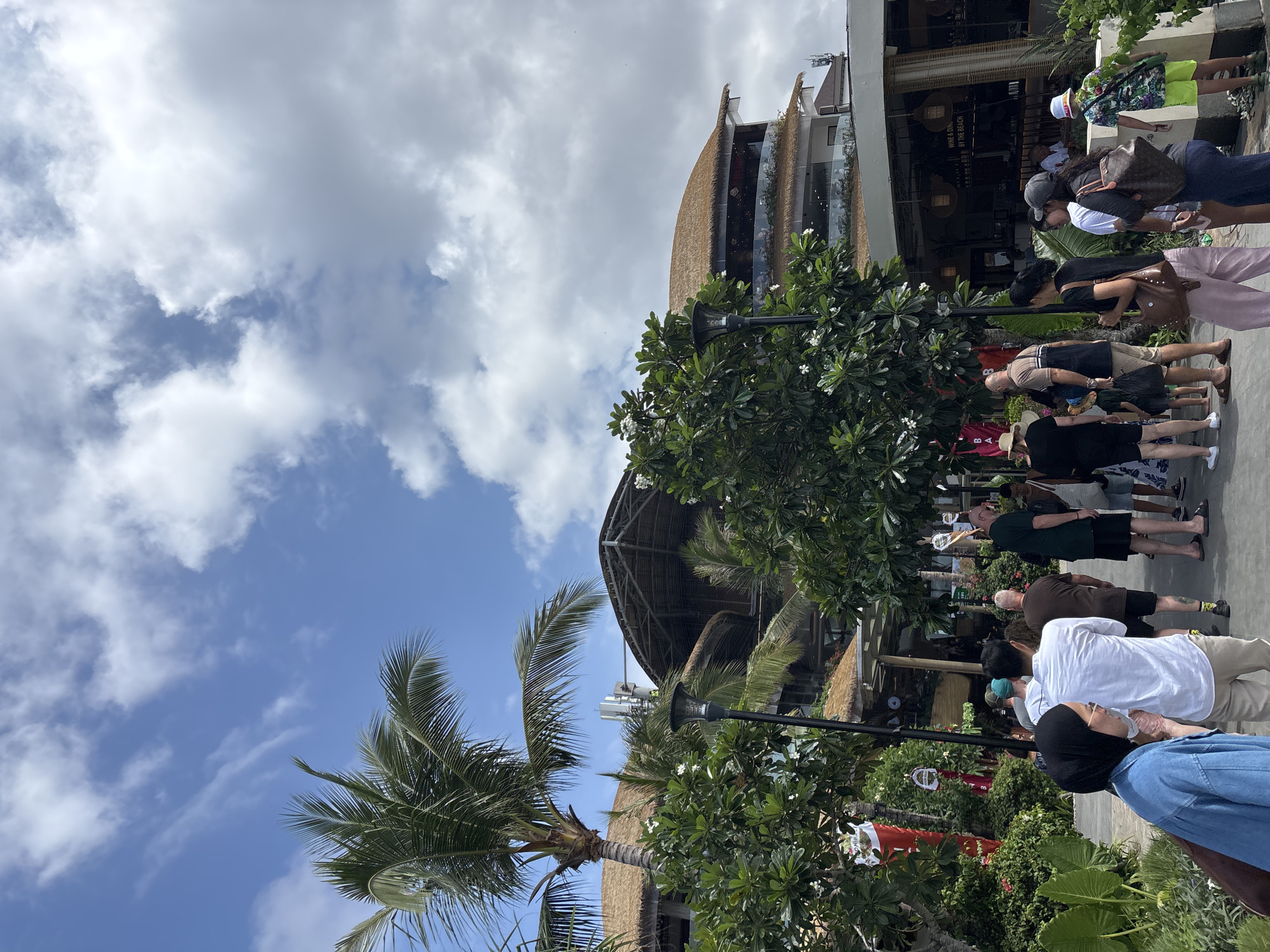 The image depicts a scene in what appears to be a tourist area in Denpasar, Indonesia, likely near a beach. A line of people, seemingly tourists, is waiting outside a building. The architecture of the building features a thatched roof and modern glass elements. Lush tropical vegetation, including palm trees and flowering bushes, is present. The sky is partly cloudy with patches of blue sky visible. There are street lamps along the street. The image is rotated 90 degrees clockwise.