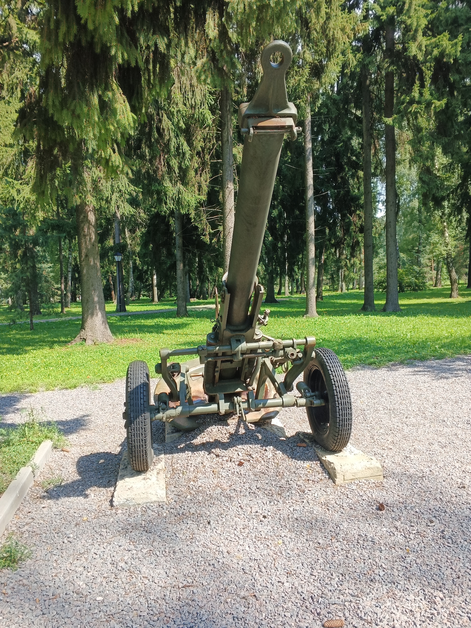 An outdoor, eye-level, full shot shows a weathered, olive-green, towed mortar positioned on a gravel surface in front of a dense forest. The mortar, elevated on two concrete blocks, faces left, its barrel angled upwards. The barrel is supported by a complex system of metal arms and wheels, with two black tires visible. In the background, tall, mature trees with thick, green foliage dominate the scene, with a well-maintained grassy area interspersed between them. Sunlight filters through the leaves, casting dappled shadows on the ground. A lamppost is visible in the middle ground. The overall impression is one of a military relic displayed in a park-like setting.