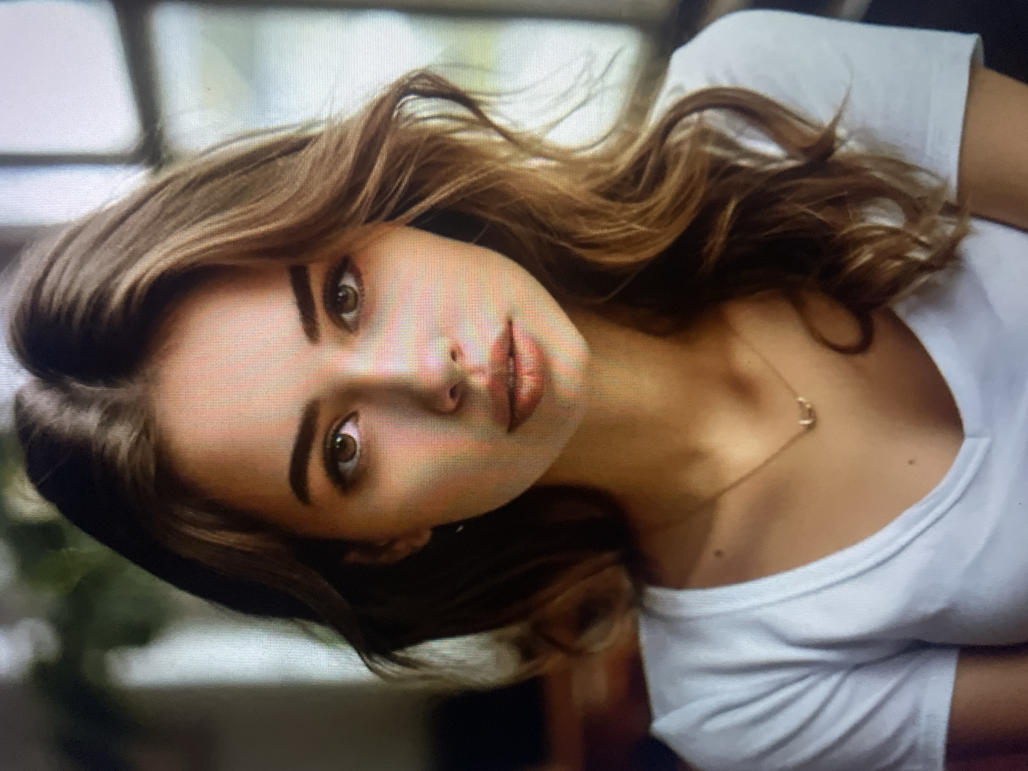 The image shows a close-up portrait of a young woman with long, wavy brown hair. She has fair skin, green eyes, and is wearing a white V-neck t-shirt and a delicate gold necklace. The background is softly blurred, suggesting an indoor setting with natural light coming from a window. There is no indication of the city of Kołobrzeg, Poland, or any urban environment. The scene is static and focuses solely on the woman's face and upper body.