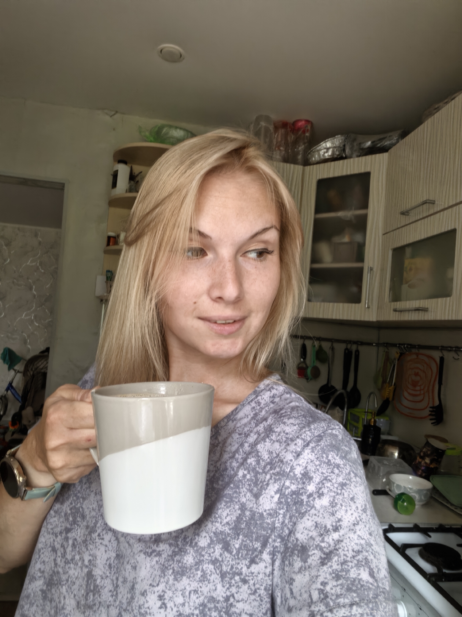 A young Caucasian woman with blonde hair and freckles is holding a white and gray mug of coffee in her left hand. She is wearing a gray t-shirt with a patterned print and a smartwatch on her left wrist. The woman is standing in a kitchen with light-colored cabinets and shelves stocked with various items, including dishes and containers. The overall environment appears to be a domestic kitchen setting. The image's metadata does not specify a location within Sterlitamak, Russia.