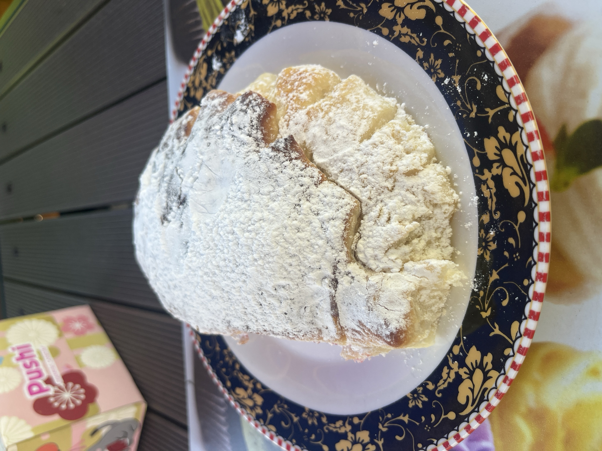 A close-up, eye-level shot shows a pastry dusted with powdered sugar on a decorative plate. The pastry appears to be a sweet bread or cake, with visible layers and a golden-brown baked exterior peeking through the white sugar. The plate has a dark blue rim with intricate gold floral patterns and a red and white striped border. In the blurred background, wooden planks suggest an outdoor setting, possibly a table. To the left, a pink and yellow box with "Pushi" written in pink lettering is also out of focus. The overall scene evokes a casual, possibly outdoor dining experience in Dzhubga, Russia.