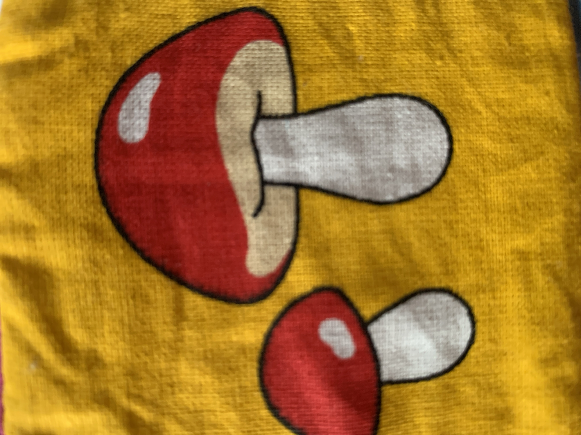 The image shows a close-up view of a yellow fabric with a pattern of two cartoon mushrooms. The mushrooms are red with white spots on their caps and white stems. They are outlined in black. The larger mushroom is on the left, and a smaller one is below and to the right of it. The fabric has a textured appearance, suggesting it could be a tea towel, placemat, or part of a garment. The overall impression is playful and decorative. No people, text, or environmental cues are visible. The setting is indeterminate.