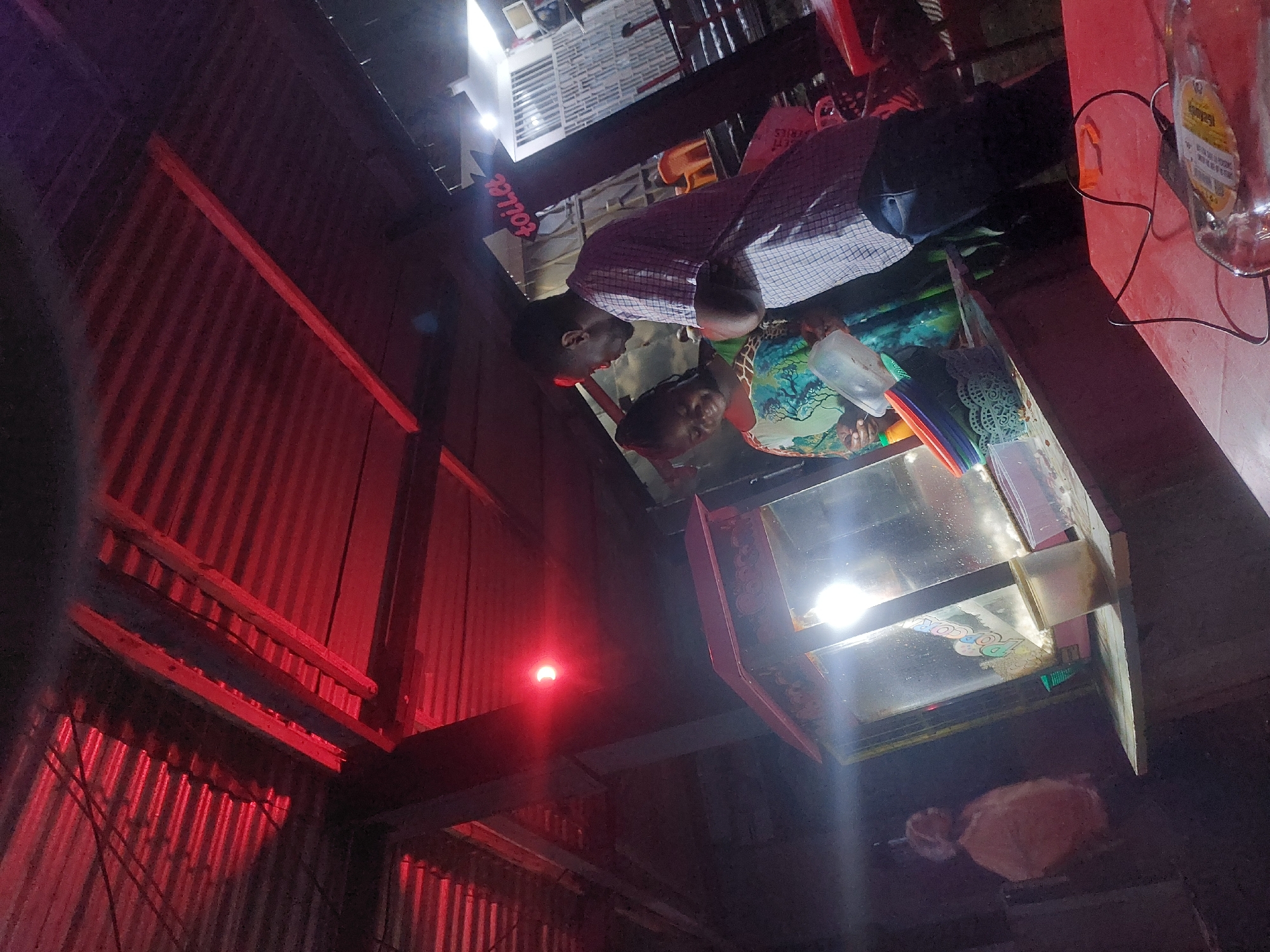 A street food vending scene in Dar es-Salaam, Tanzania, at night.
Two people, likely vendors, are standing by a glass-fronted food cart. One is an adult male wearing a checkered shirt. A second person, likely female, is nearby. The cart appears to be selling popcorn as the word "Popcorn" can be seen on the front. Various items are arranged on the cart's surface.
The backdrop reveals a corrugated metal structure illuminated by red light. A beam crosses the top of the image frame, and above it are partially visible structures. A light source is present in the dark area below the vending cart. A bottle hangs near the top right corner of the image. Electrical wires are visible along the side.
