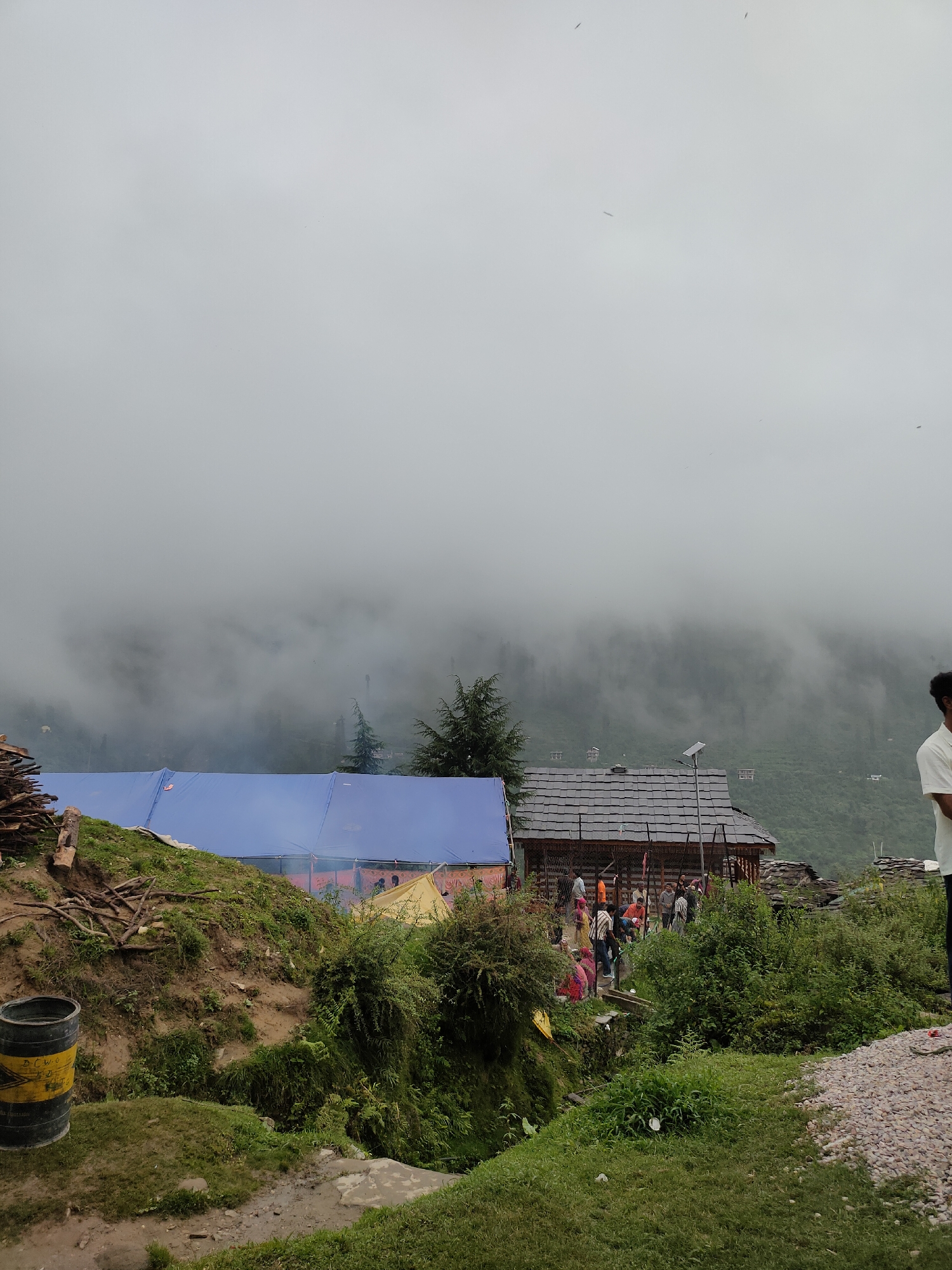 The image depicts a cloudy day in Damadi, India, with a focus on a village settlement nestled against a misty hillside. In the foreground, a large blue tarp stretches across a grassy slope, suggesting a temporary shelter or event setup. To the left, a pile of logs and branches lies near a yellow and black barrel.
A traditional wooden building with a dark, multi-layered roof sits prominently in the mid-ground. Several people are gathered around and in front of this building, some appearing to be engaged in conversation or activity. A solar panel is mounted on a pole near the building, indicating a source of power.
The background is dominated by a steep, green hillside, partially obscured by thick white clouds and mist that cling to the trees and landscape, creating a diffused and atmospheric scene. Several small structures are visible on the distant slopes, suggesting the presence of more dwellings or a wider village area. On the far right, a person in a white shirt stands partially visible, facing away from the camera. The overall impression is of a remote, possibly rural, community experiencing overcast weather.