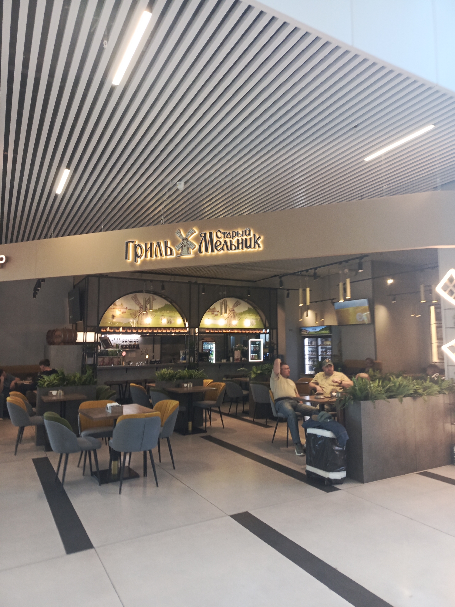 The image depicts the interior of a cafe or restaurant with the signage "Гриль Старый Мельник" (Grill Old Miller) prominently displayed. The cafe has a modern design with a slatted ceiling and ambient lighting. Several tables and chairs are arranged throughout the space, with some patrons seated at them. In the foreground, there are four chairs around a table, and further back, two men are seated at a table, appearing to be engaged in conversation or waiting for their order. One man is leaning back in his chair with his arm raised, while the other is sitting upright. A large black trash bag is placed on wheels next to the seating area. The overall atmosphere suggests a casual dining environment. The location is specified as the city of Ob, Russia.