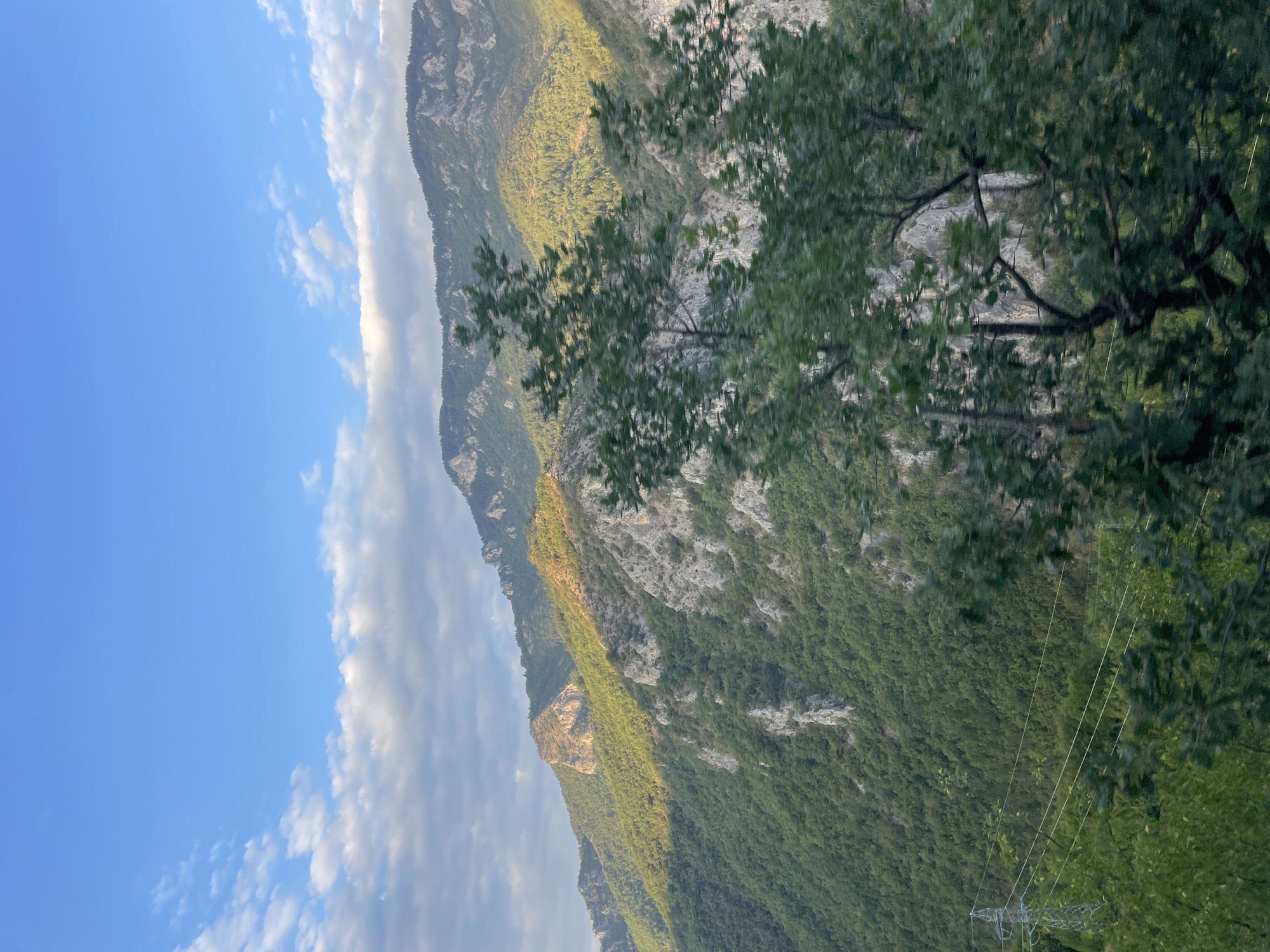 This is a landscape photograph taken in the city of Yugovo, Bulgaria. The image depicts a mountainous region with lush green forests covering the slopes. Rocky outcrops are visible on the mountainsides, and the peaks are silhouetted against a clear blue sky dotted with white clouds. The sun is casting a warm glow on some parts of the mountains, highlighting the textures and contours of the terrain. In the foreground, there are trees with green foliage, some of which are slightly blurred due to movement. A power line tower with wires running across can be seen on the left side of the image, amidst the greenery. The scene conveys a sense of natural beauty and tranquility.