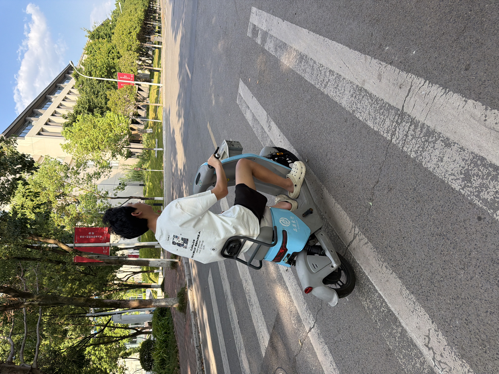 A young person, viewed from behind, rides a light blue and gray electric scooter across a pedestrian crossing. The person is wearing a white t-shirt with a graphic print on the back and dark shorts, along with beige clogs. The scooter has a small basket in front and a black metal frame behind the seat. The scene takes place on a paved road with white pedestrian crossing lines. In the background, there are trees with lush green foliage, and a modern building with a beige facade and many windows. Red banners hang from poles along the sidewalk. The sky is blue with scattered white clouds, indicating it is daytime. This scene is located in the city of Jiangjin, China.