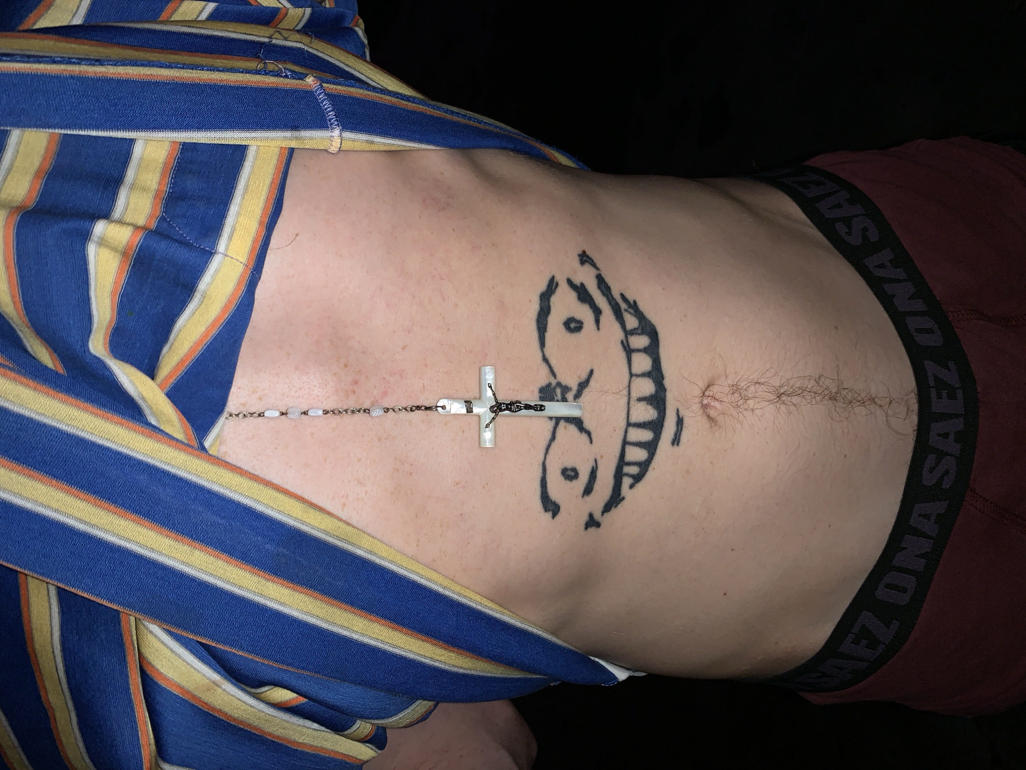 The image shows a close-up of a person's upper torso and midsection, likely taken indoors in a dimly lit or dark room at night, with a black background surrounding the subject. The person is lying down, wearing a blue, yellow, and orange striped long-sleeved garment that is partially pulled up, revealing the abdomen. Beneath this, a maroon pair of underwear is visible, with the black elastic waistband displaying the brand name "ONA SAEZ" in light-colored text.
Draped across the person's upper chest and stomach is a rosary-style necklace with small white or light-colored beads. It features a prominent white cross with a metallic crucifix figure attached. On the left side of the upper abdomen, there is a large, black tattoo depicting a stylized, smiling face with wide eyes and an open mouth. The skin has a fair complexion with some visible freckles, and there is light body hair around the navel area. The overall scene is a candid shot of the person in a relaxed state in what appears to be a private setting in Buenos Aires, Argentina.