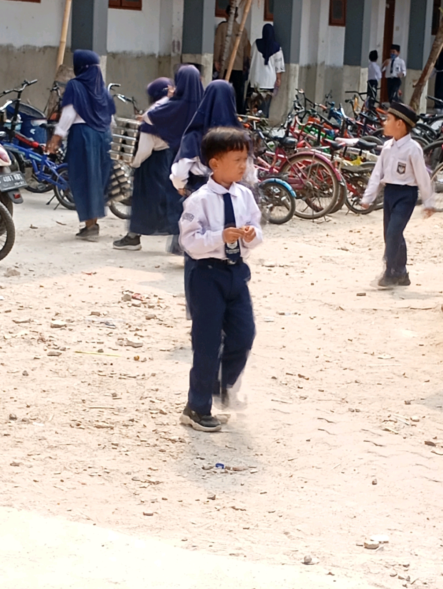 In Babelan, Indonesia, a young boy in a white shirt and dark blue pants, wearing a small dark cap, walks on a dirt path. He appears to be holding a small item in his hands. Several other children, mostly girls in school uniforms consisting of headscarves and long skirts, are visible in the background, some standing near bicycles parked in front of a building. The environment suggests a schoolyard or a communal area with parked bicycles and a sandy, uneven ground.