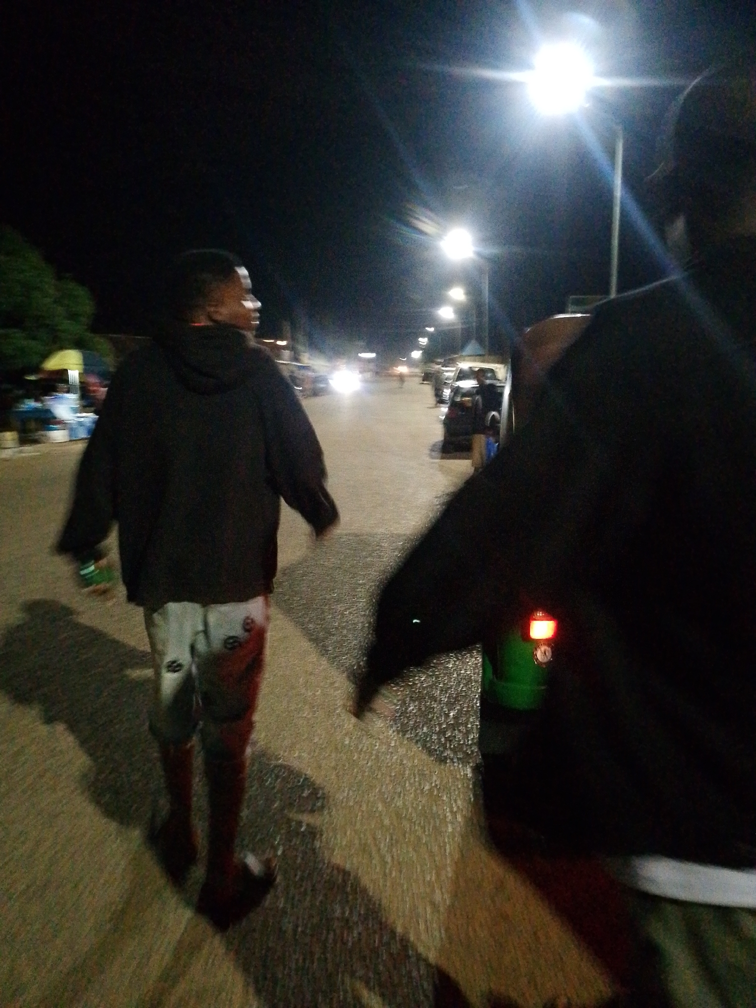 A person walks at night on an asphalt road with streetlights and some trees visible. The person is wearing a black hoodie and light-colored shorts with red markings. Another person is partially visible in the foreground on the right, also at night. The scene appears to be in an urban or suburban area.