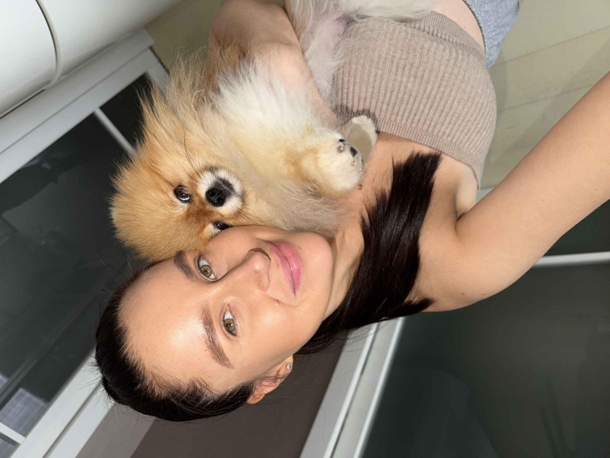 A light-skinned female with dark hair is holding a light brown and white Pomeranian dog. The woman is wearing a tan top and grey sweatpants. She has green eyes and is wearing earrings. The dog is laying on her shoulder. The background includes a white refrigerator, grey walls, and a partial view of a window. The scene may be located in an apartment or house. There is no information in the image to confirm the location as Khimki, Russia.