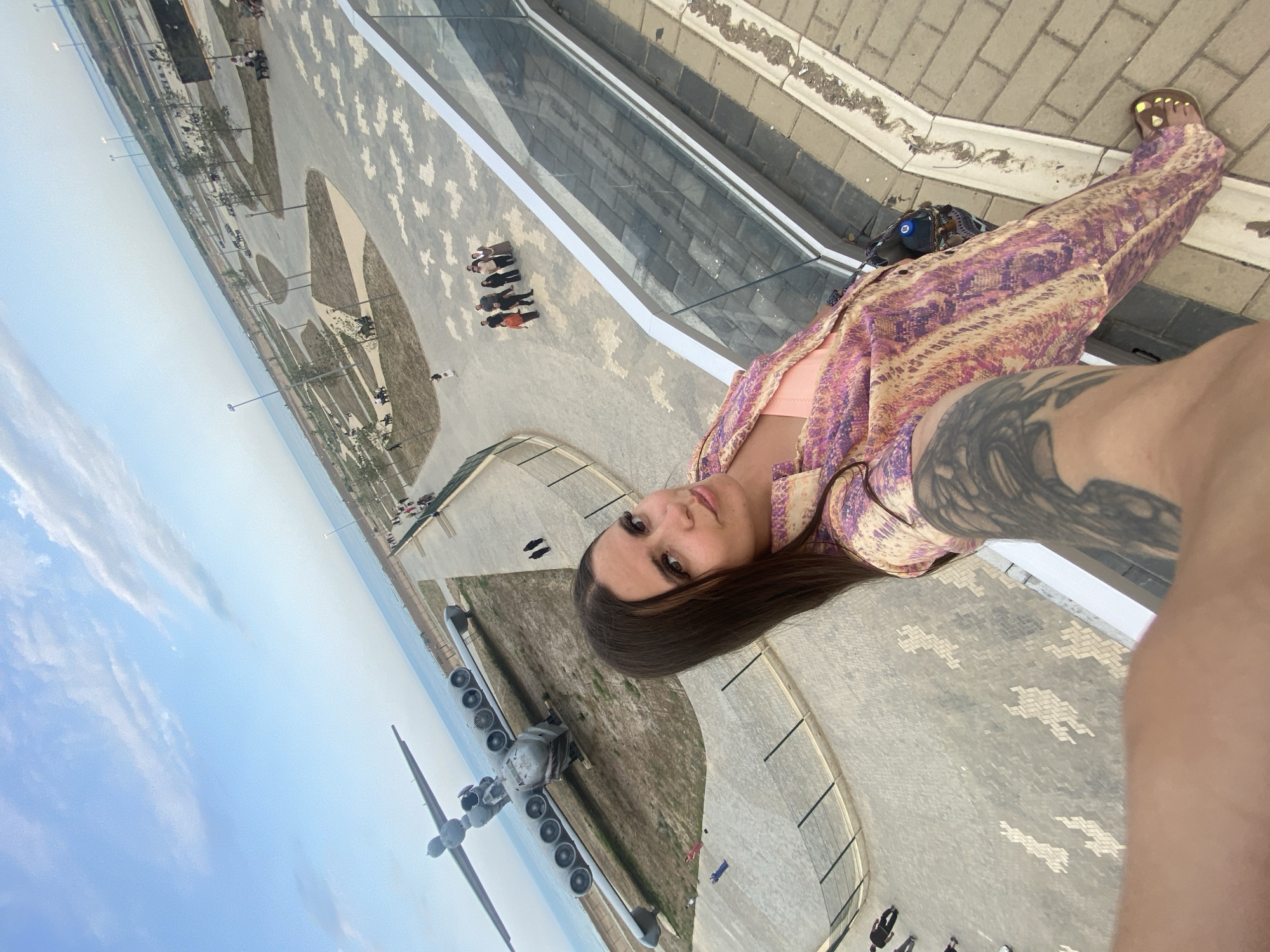 A high-angle, wide shot captures a woman in the foreground, her arm extending into the frame holding the camera. She is wearing a vibrant, patterned outfit in shades of pink, purple, and yellow, and appears to be standing on a paved area with a glass railing. Behind her, a large, gray airplane rests on a grassy, dry field, its numerous engines visible. The plane is positioned near a body of water, likely the Caspian Sea, given the presumed location of Arablyar, Russia. Beyond the plane, a park area with walking paths, benches, and scattered trees is visible, with a group of people gathered in the middle distance. The sky is a clear blue with sparse, wispy clouds. The overall scene suggests an outdoor exhibition or park setting with a historical aircraft as a centerpiece.