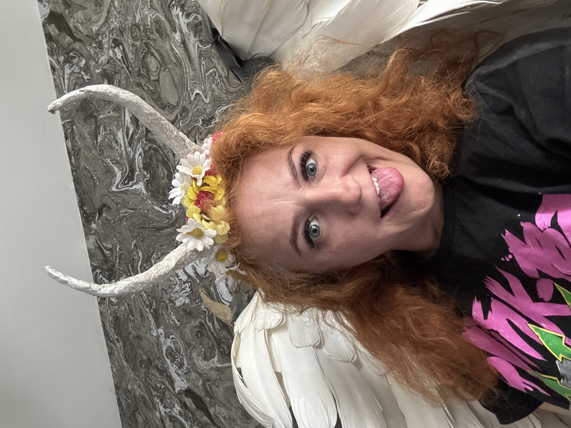 A woman with red hair, blue eyes, and a whimsical costume is photographed indoors. She is wearing a black shirt with a large, stylized pink and green design. She has a headpiece with white horns decorated with white and yellow flowers. She also has large white wings attached to her back. The woman is sticking her tongue out and looking playfully at the camera. The background is a grey and white marbled wall. The image was taken indoors and the location is the city of Chaikovsky, Russia.