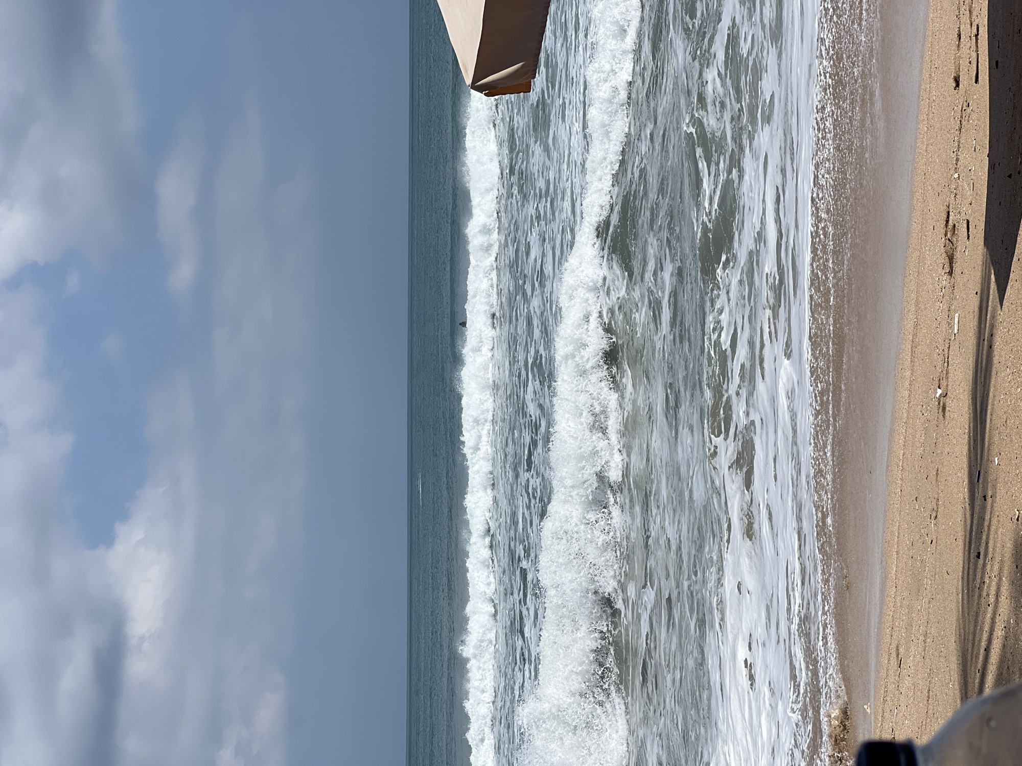 The image depicts a beach scene in Kuta, Indonesia, with a focus on the ocean and waves. In the distance, a solitary surfer is visible in the turquoise water. The foreground shows a sandy beach with some debris, and a portion of a tan beach umbrella is visible on the right side of the frame. The sky above is partly cloudy with a mix of blue sky and grey clouds. The waves are crashing onto the shore, creating white foam and splashes.