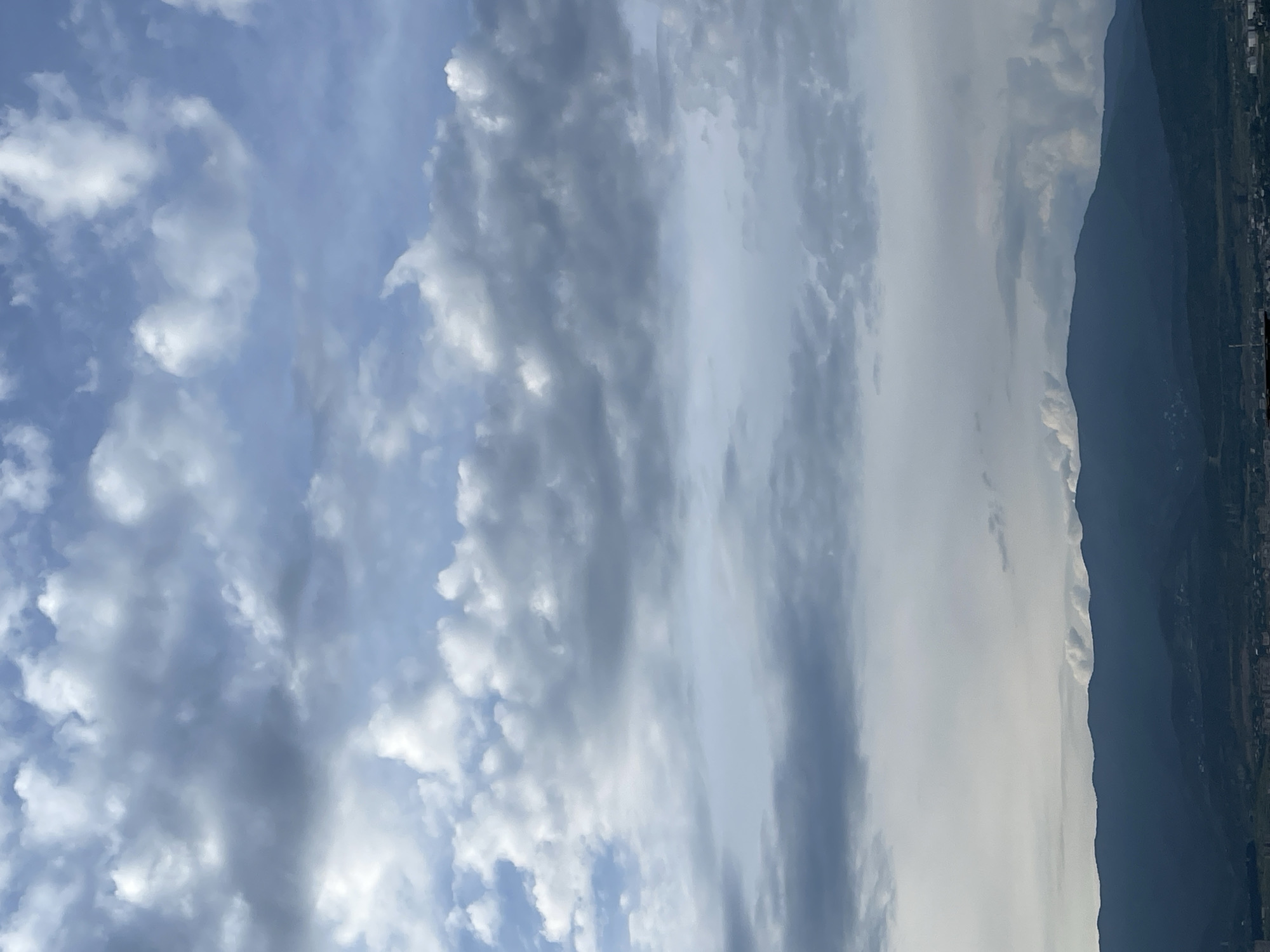 The image depicts a panoramic view of the landscape surrounding Tbilisi, Georgia, on what appears to be an overcast day with scattered clouds. The foreground is dominated by the dark, undulating silhouette of a mountain range, likely the foothills of the Trialeti Range, which encircles the city. Subtle details of a settlement, possibly a suburb or village, are visible on the slopes and in the valleys between the mountains, characterized by small clusters of buildings and hints of infrastructure. The sky above is a dynamic canvas of clouds; the lower portion features darker, heavier cloud formations that transition into lighter, brighter clouds towards the upper half of the image. There are no people or animals visible in the frame. The overall scene conveys a sense of natural grandeur juxtaposed with human habitation at the edge of a mountainous region.