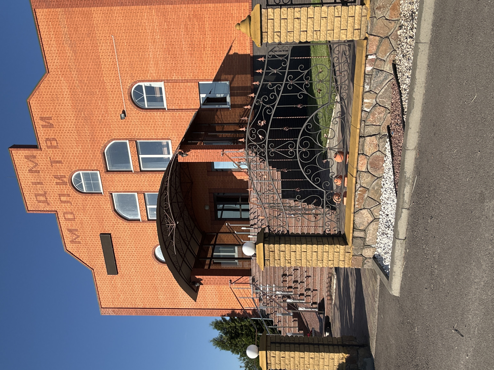 The image shows a bright orange brick building with the words "ДІМ МОЛИТВИ" (House of Prayer) inscribed in dark letters on its facade under a clear blue sky. The building features several arched windows and a covered entrance with a metal awning leading to a set of stone stairs. A decorative metal fence with spherical lights on stone posts stands in front of the building, separating it from a paved area and a lawn. The setting appears to be a peaceful, well-maintained exterior, possibly in the village of Dovhe, Ukraine, given the context.