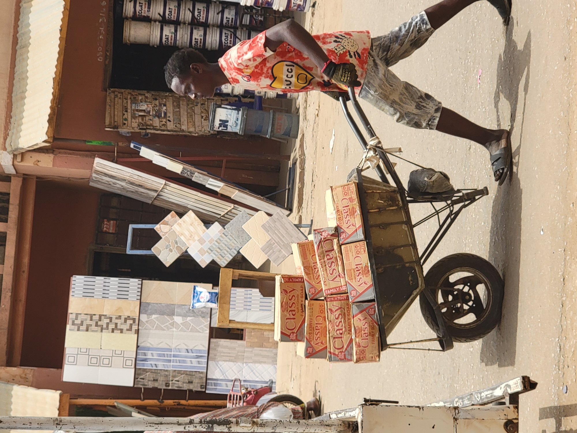 A man pulls a cart loaded with "Classy" family soap boxes down a street in Maiburiji, Nigeria. The cart is a metal, single-wheeled design with a filled hat sitting on top. The man, who is wearing a printed t-shirt, camouflage shorts, and sandals, walks along a sunlit, paved surface. In the background, there are paint cans and ceramic tiles displayed outside a storefront.