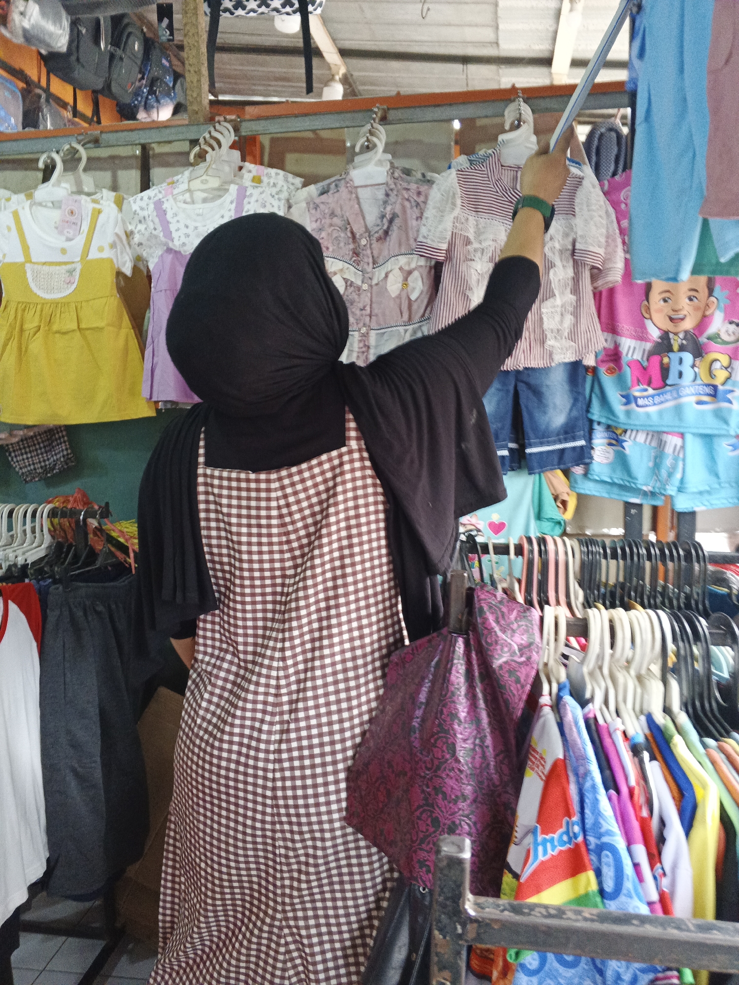 The image captures a street stall in Warungkiara, Indonesia, showcasing an array of children's clothing. A person wearing a black headscarf and a checkered apron stands with their back to the camera, holding a long pole, possibly to adjust the hanging garments. The stall features racks of colorful clothes, including dresses, shirts, and shorts, with various patterns and designs. The overall impression is one of a busy marketplace, with goods neatly displayed for potential customers.