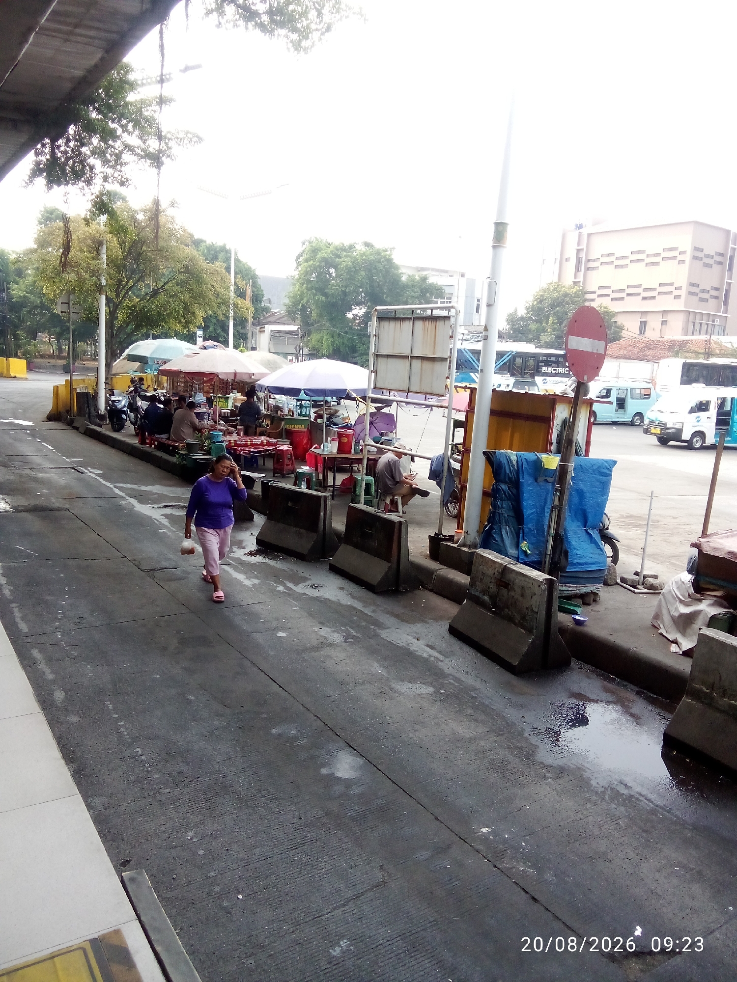 The image depicts an outdoor market scene in East Jakarta, Indonesia, likely on August 20, 2026, at 09:23. A woman in a purple shirt and pink pants walks on a wet asphalt road, holding a clear plastic bag. She is facing left and appears to be touching her ear or hair.
In the background, several market stalls are set up with umbrellas providing shade. People are visible sitting and standing at these stalls, some interacting with vendors. Motorcycles are parked behind the stalls.
Further in the background, various vehicles are present, including buses and minivans. A red and white "no entry" sign is prominently displayed on a pole. The environment appears to be a roadside area with concrete barriers along the edge of the market. Buildings and trees are visible in the distance. The ground is wet, suggesting recent rain.