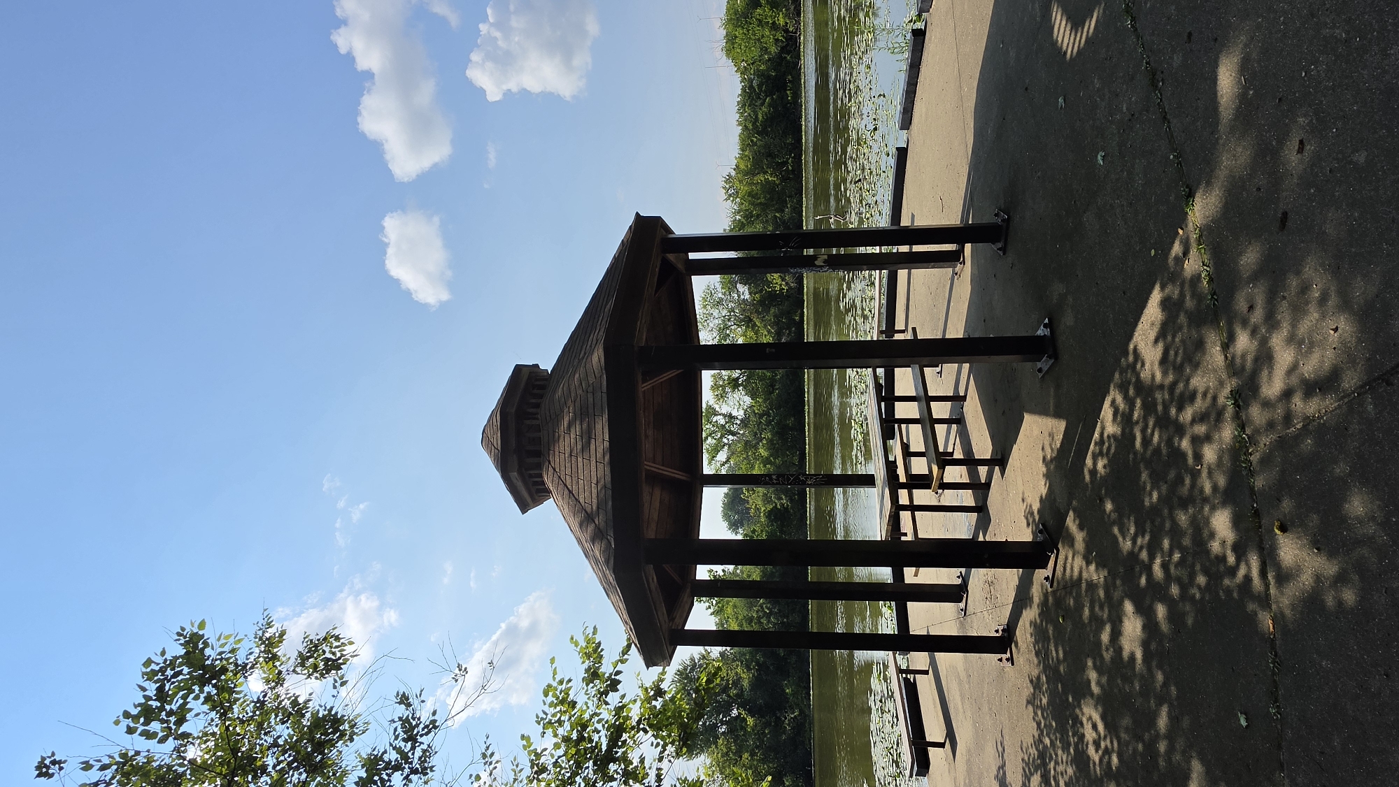 The image shows a hexagonal wooden gazebo with a brown shingled roof, situated on a concrete platform next to a body of water. Inside the gazebo are two wooden picnic tables with benches. To the left and right of the gazebo are additional benches. The water, likely a lake or river, is covered in lily pads and bordered by a dense line of green trees. The sky is a clear blue with scattered white clouds. The scene is illuminated by sunlight, casting dappled shadows from nearby trees onto the concrete platform. The location is identified as Elgin, United States of America, suggesting a public park or recreational area. There are no people visible in the image.