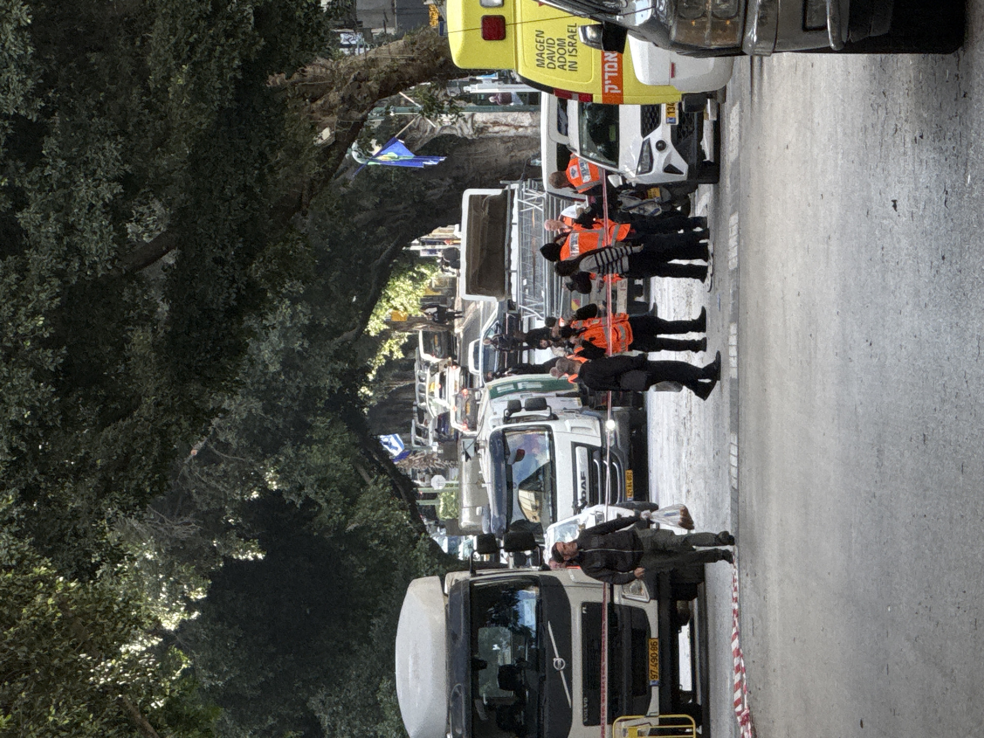 The image depicts a street scene in Ramat Gan, Israel. A line of trucks and vehicles are stationary on the road, with a group of individuals wearing orange vests standing alongside them. A man in a dark jacket is also standing near the front of the line of trucks. A yellow ambulance is visible near the front of the line. The street is flanked by trees and foliage.