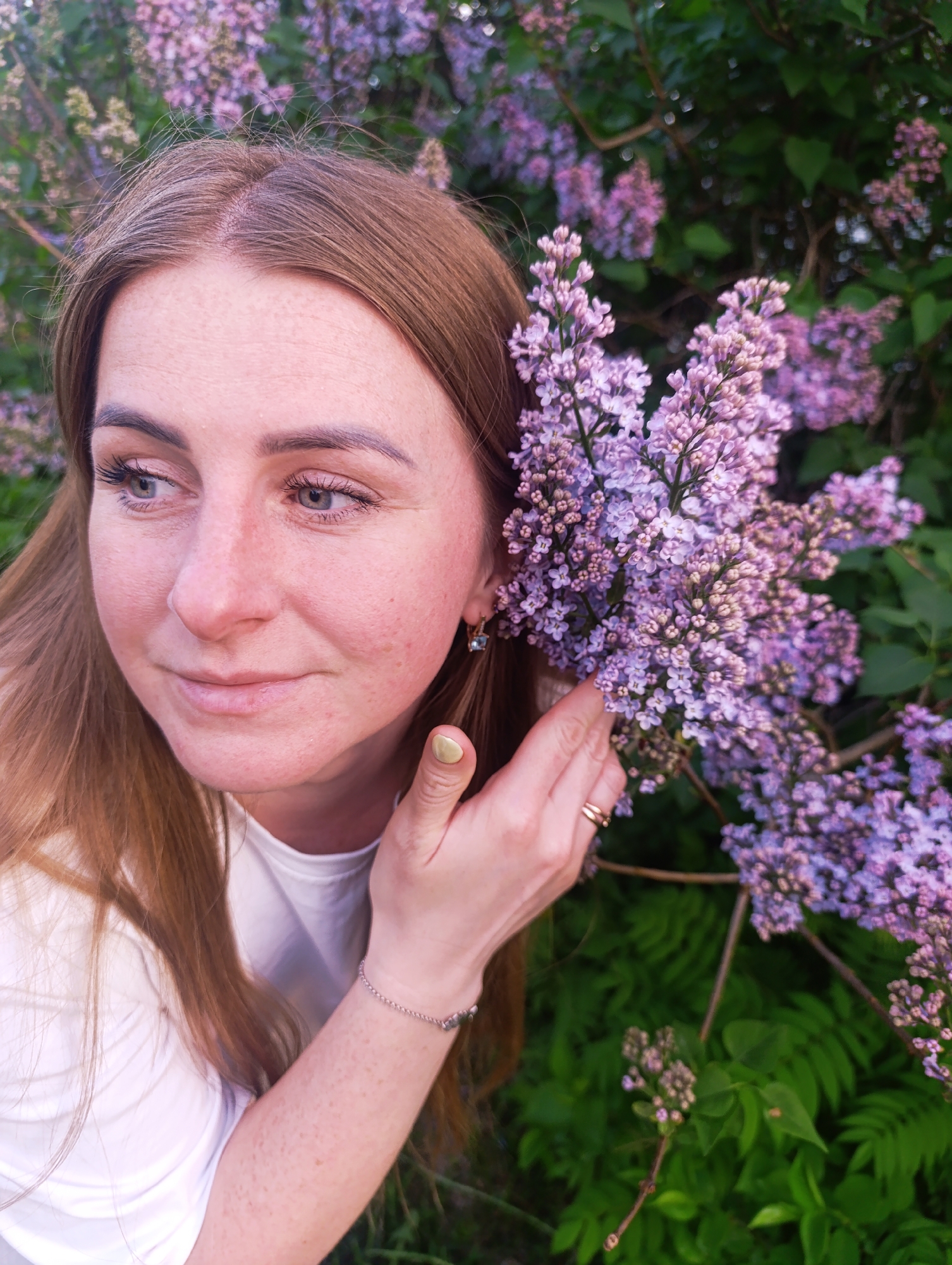 A woman with light skin, freckles, and light brown hair stands near a lilac bush. She is wearing a white t-shirt and an earring. Her hand, adorned with a gold ring, is gently placed near the lilac blossoms. The lilac bush, with its purple flowers and green leaves, fills the background. The location is Unknown, Unknown.