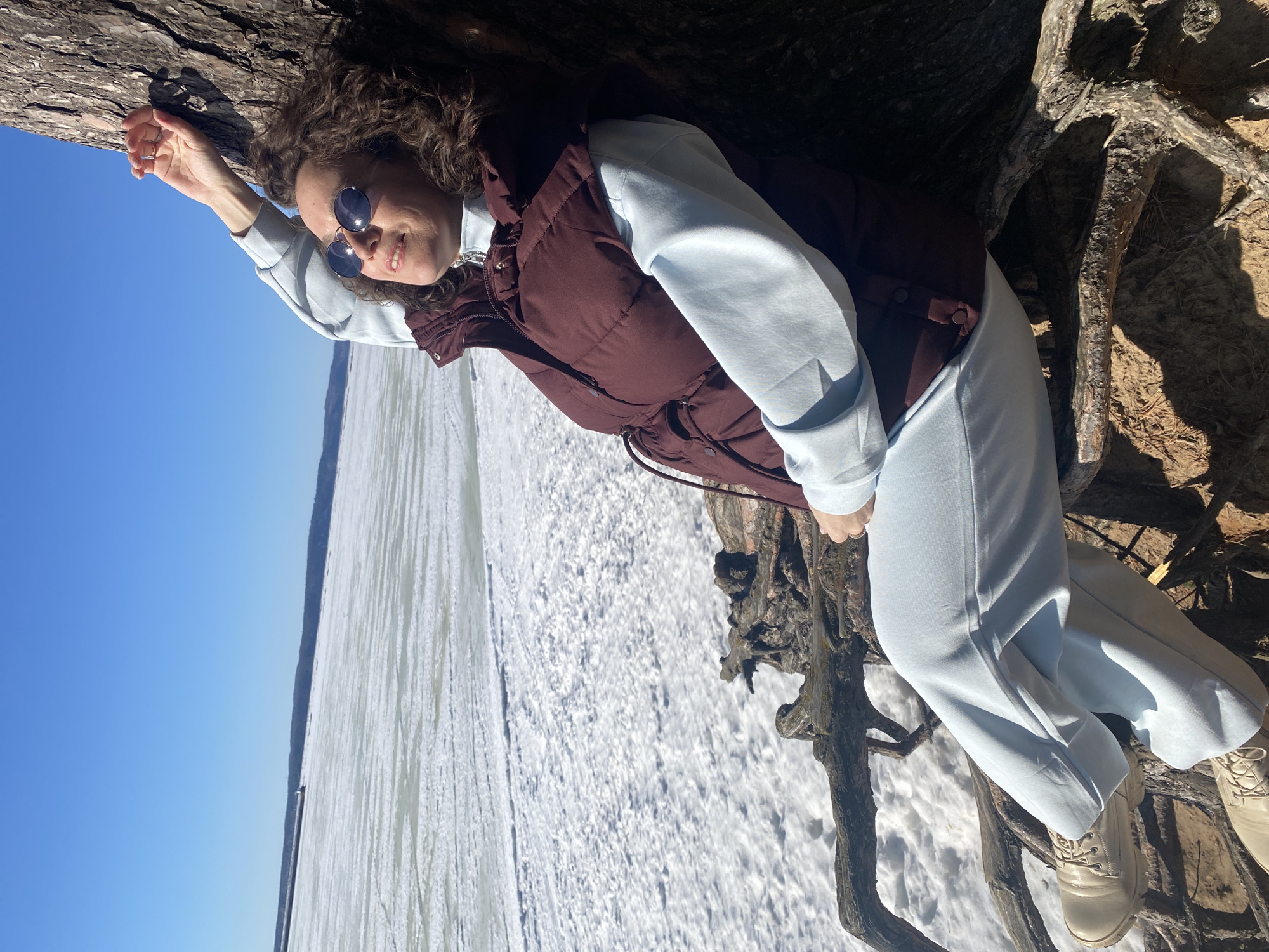 An adult female is depicted seated against the exposed roots and trunk of a large tree on a snow-covered embankment. The scene overlooks a vast frozen body of water, identified as the Votkinsk Reservoir near Chaikovsky, Russia. The individual has curly brown hair and wears dark, circular sunglasses, a light blue quarter-zip long-sleeved top, a dark maroon sleeveless puffer vest, light blue trousers, and light-colored lace-up boots. Her left arm is raised and rests against the tree trunk, while her right hand rests near her midsection. She is looking upwards with a slight smile. The immediate ground around the tree consists of exposed soil and patches of white snow. Beyond the embankment, the frozen reservoir is covered in a layer of snow and ice, displaying some textural variations. A dark, forested shoreline is visible on the distant horizon, with a small, light-colored structure, possibly a navigational aid or building, discernible on the far bank. The sky above is clear and bright blue. The scene is illuminated by strong, direct sunlight, casting distinct shadows.