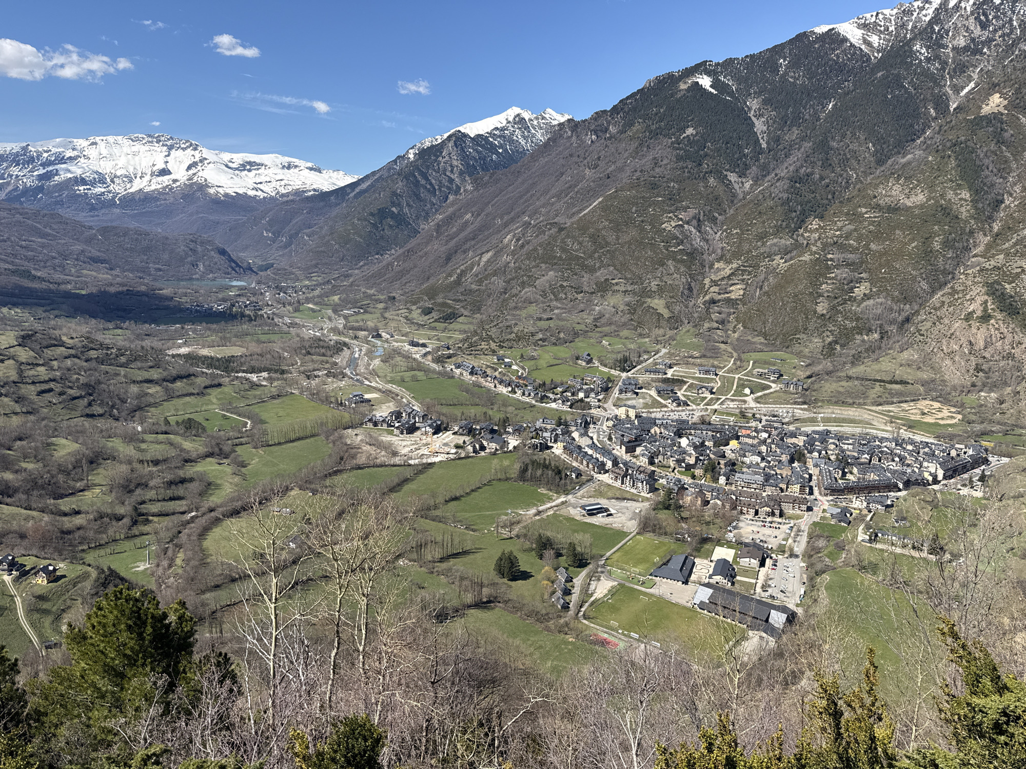 The image is a high-angle, wide-shot photograph of a town nestled in a valley surrounded by mountains, some of which are snow-capped. The sky is clear and blue.
The town, presumably Benasque, Spain, is located in the center-right foreground. It is densely built with buildings varying in size, most with dark roofs. The buildings appear to be a mix of residential and commercial structures. In the foreground, there are some modern, low-rise buildings with parking areas and sports fields.
The valley is a mix of green fields and patches of forest. A river or stream runs through the valley. In the distance, there is a body of water that could be a lake or reservoir.
The mountains rise steeply on both sides of the valley. Some of the peaks are covered with snow. The slopes of the mountains are mostly covered with trees. The terrain is rugged and steep.
The foreground shows vegetation with trees and shrubs. They are mostly bare, indicating it may be early spring. The overall scene is one of a picturesque town in a stunning mountain setting.