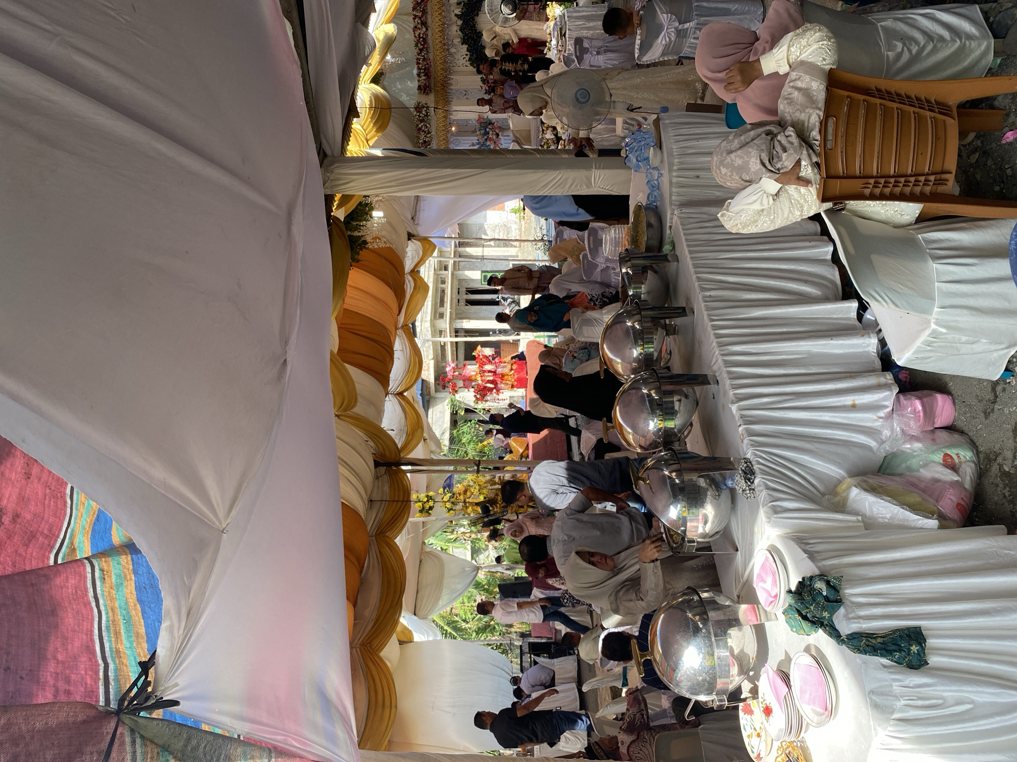 This image captures an outdoor event, likely a wedding reception, under a colorful tent adorned with yellow and white fabric. The scene unfolds in Payaudeung, Indonesia. A long table laden with food in silver chafing dishes dominates the foreground, set with plates and pink napkins. Several people are visible, some mingling, others seated and observing. In the center, a woman in a beige hijab attends to the food. To the right, two women in hijabs are seated, one appearing relaxed, leaning back in a plastic chair. The background reveals more seating areas, a stage with floral decorations, and onlookers. The environment is a courtyard, with a dirt ground and the suggestion of buildings and vegetation beyond the tent.