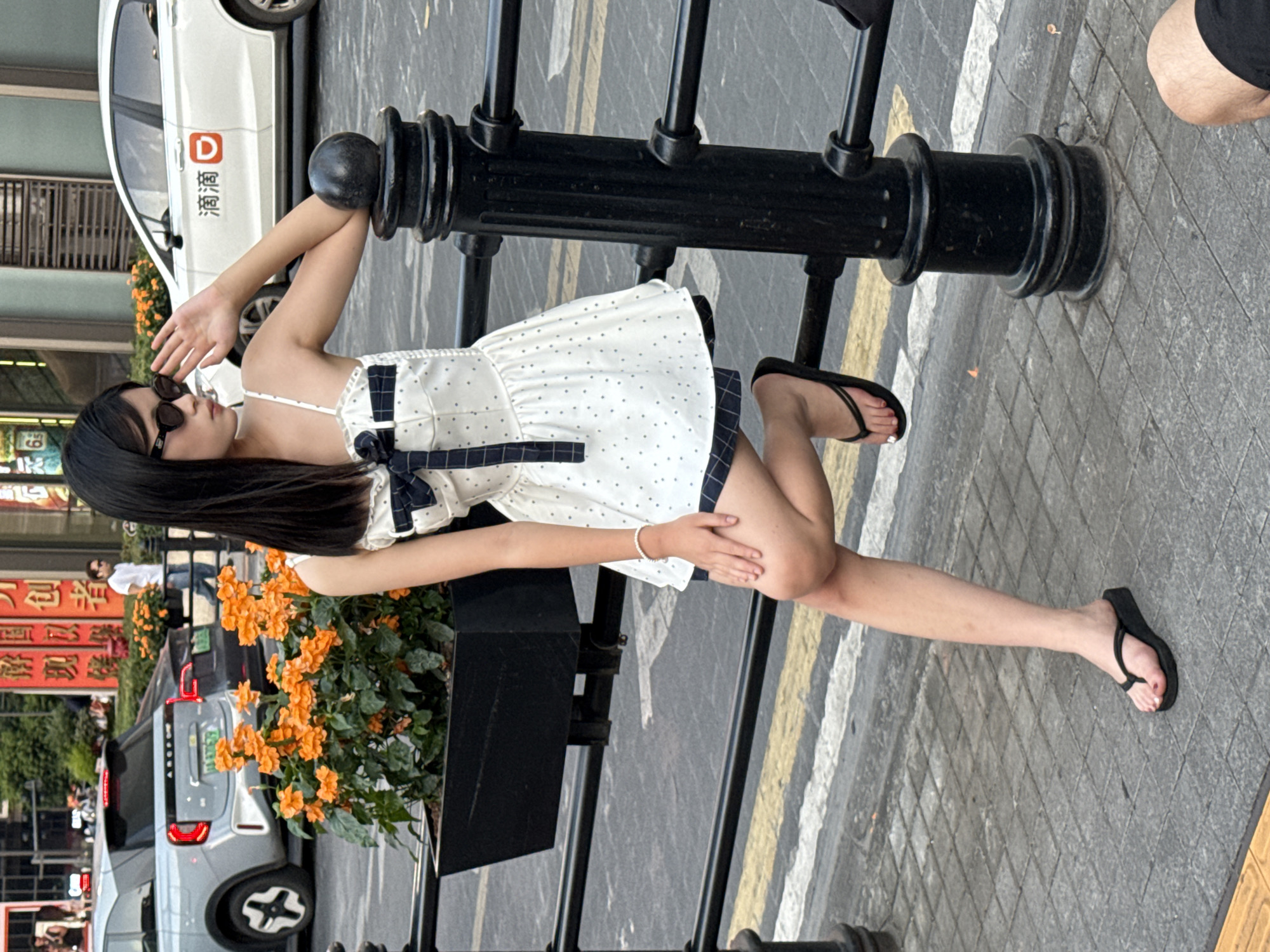 A young East Asian woman with long black hair and sunglasses poses on a sidewalk in Yuzhong District, China. She is wearing a white polka dot dress with a navy blue ribbon tie at the bust and a navy blue plaid trim at the hem. She has a pearl bracelet on her left wrist and is wearing black flip-flops. She is leaning against a black metal fence post, with her left leg bent and resting on the fence. Behind her, there is a planter box filled with bright orange flowers. In the background, there are cars on a street, including a white car with Chinese characters and a red logo, and a silver car. The sidewalk is paved with grey square tiles and marked with yellow and white lines.