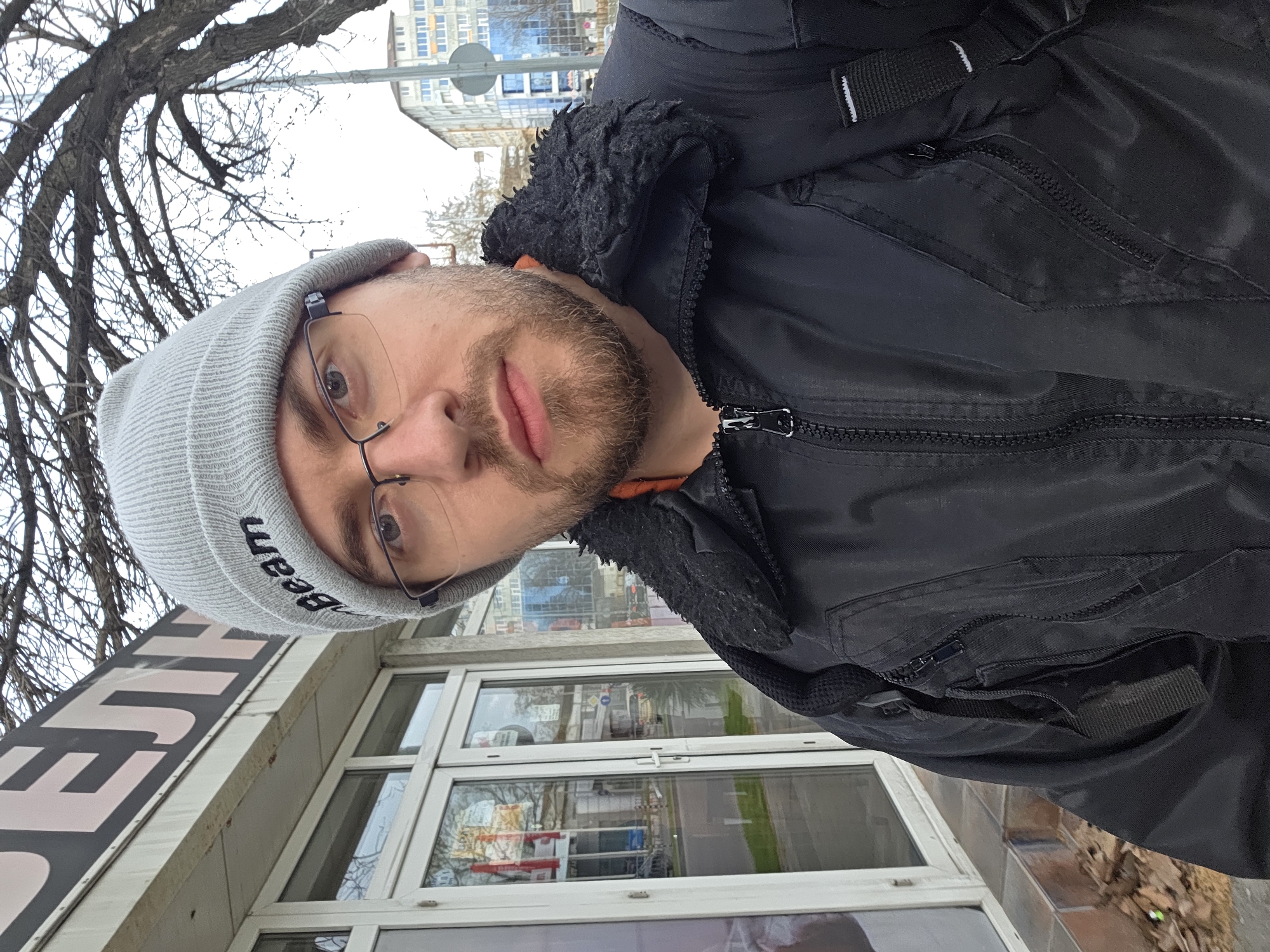 A man wearing a grey beanie with the word "Deem" on it and glasses is standing outside a building. He's wearing a black jacket with a furry hood. The background suggests an urban environment. The building behind him has large windows. Above the building, there is a bare tree and a taller building in the distance. Based on the architecture and the Cyrillic-script signage visible in some reflections, the location is likely Plovdiv, Bulgaria.