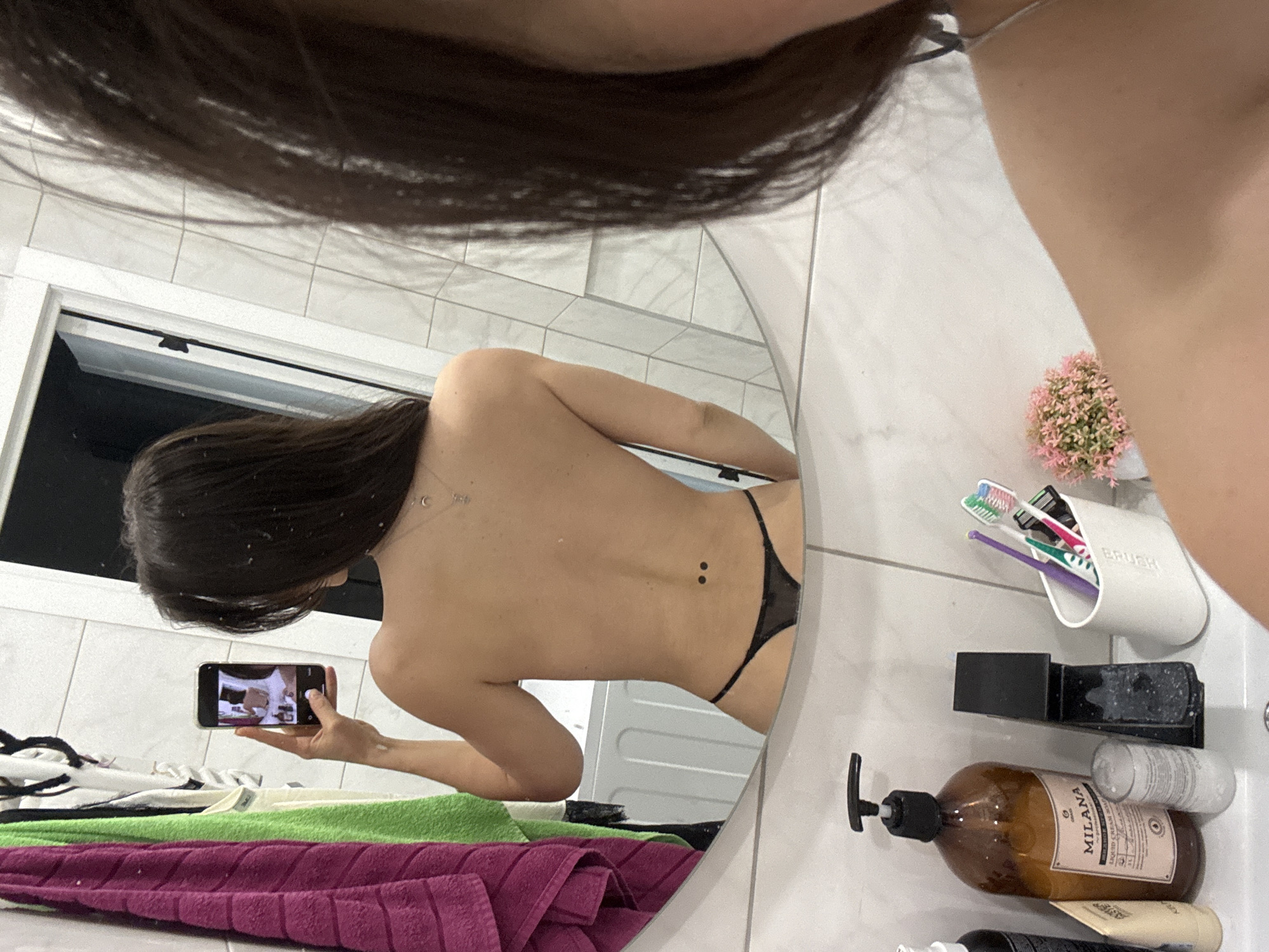 The image provides an overhead perspective of a bathroom, partially revealing the dark hair and arm of the person holding the camera. The primary subject is a woman, reflected from behind in a large, round mirror. She has long, dark hair and is wearing a black thong, appearing bare-chested.
The woman in the reflection is actively holding a black smartphone horizontally with both hands, pointing it towards her lower body, suggesting she is taking a picture or video of herself in the mirror. Her phone's screen shows a bright, somewhat indistinct image. Visible on her lower back are two small, dark dermal piercings, and a delicate silver necklace chain can be seen around her neck/upper back area.
The bathroom features white tiled walls and floor. On the sink counter to the right of the mirror, various toiletries are organized. These include a prominent brown pump bottle labeled "MILANA LIQUID HAND SOAP," a white holder inscribed with "BRUSH" containing multiple toothbrushes (with green, purple, white, and blue bristles) and a tube of toothpaste. A small pot of pink artificial flowers, a black holder, and other smaller bottles are also present. Below the mirror, a stack of colorful folded towels in white, green, and dark pink/maroon is visible. A white door frame is reflected in the upper left of the mirror, suggesting another room beyond. The scene depicts a private, modern bathroom setting.