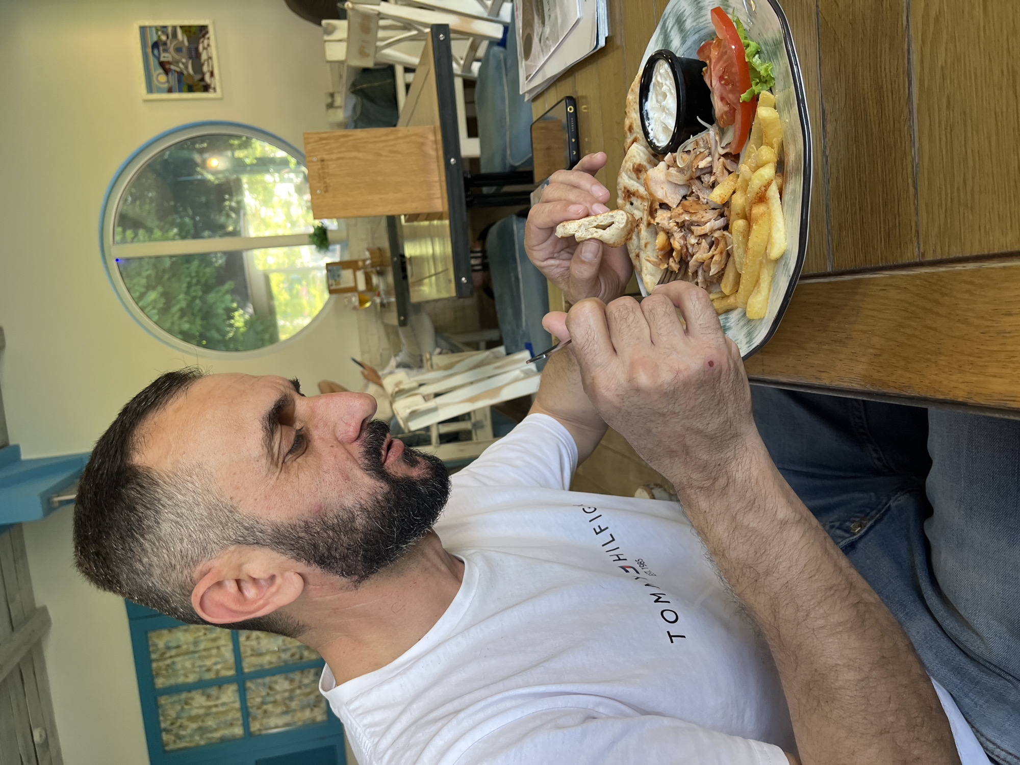 A male individual, approximately 30-50 years old, with short dark hair exhibiting some graying at the temples and a dark, full beard, is seated at a light-colored wooden table in a restaurant located in Sochi, Russia. He is wearing a white short-sleeved t-shirt with the phrase "TOMMY HILFIGER EST. 1985" printed in red and black on the chest, and blue denim jeans. His head is angled downwards, focusing on the plate in front of him. In his left hand, he holds a silver fork, while his right hand holds a small, torn piece of pita bread.
The plate, which is round and features a green and white patterned rim, contains a meal comprising shredded grilled meat, a portion of french fries, two slices of red tomato, a small quantity of green leaf lettuce, and a small black ramekin filled with a white sauce. Several pieces of pita bread are also present on the plate.
The restaurant interior features light-colored wooden tables and white chairs with light blue cushions. A black smartphone is positioned on the table to the right of the meal. In the background, a large circular window with a light blue frame offers a view of green foliage. To the right of this window, a framed painting depicting a white building with a blue dome against a sunset sky is mounted on a light-colored wall. On the left side of the frame, a rectangular window with light blue frames and patterned curtains is visible. The ceiling is light-colored with visible wooden beams. The scene depicts a casual dining experience.