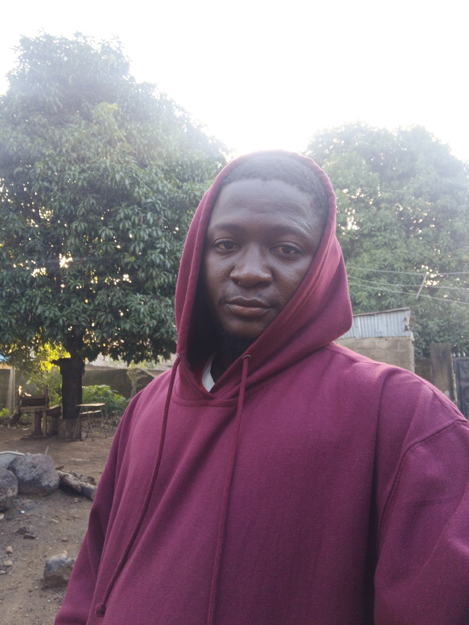 A dark-skinned adult male is depicted from the chest up, positioned centrally in an outdoor setting in Akwanga, Nigeria. He is wearing a maroon hooded sweatshirt with the hood drawn over his head, revealing a glimpse of a white collar or undershirt at the neckline. The subject has short dark hair visible beneath the hood, a full beard or stubble, and maintains a direct gaze toward the camera with a neutral expression. The background features dense green foliage from large trees, with a bright, overexposed sky above, indicating strong sunlight, possibly from behind or above. On the lower left, an earthen ground is visible with scattered rocks, and a rustic wooden table or bench is present further back. To the right, a structure featuring corrugated metal roofing and concrete block walls is partially visible behind the subject's right shoulder. The scene is illuminated by natural, bright daylight.
