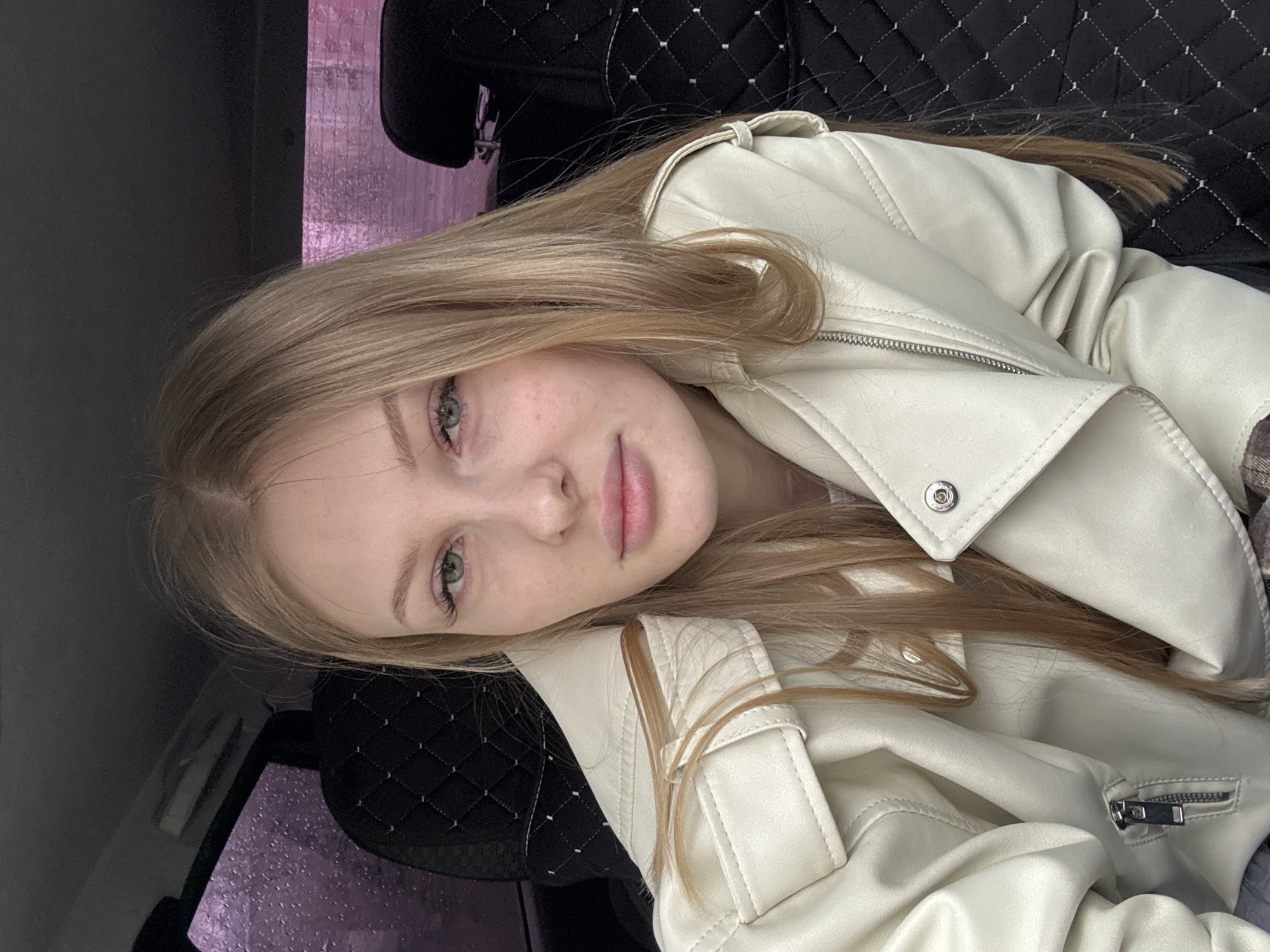 A young woman with long, straight blonde hair and light-colored eyes is captured in a close-up portrait, rotated 90 degrees counter-clockwise. She has a fair complexion with faint freckles and her lips are slightly parted, looking directly at the camera with a calm expression. She is wearing an off-white or cream-colored faux leather jacket with a prominent wide collar, zippered details, and snap buttons.
The setting appears to be the interior of a vehicle, likely a car, with black quilted seats forming a distinctive background pattern behind her head and shoulders. Through a window visible in the upper left, a blurry background with a vibrant purple or pink hue is discernible, possibly indicating artificial lighting outdoors or a reflection. Water droplets are visible on the windowpane, suggesting it might be raining or has recently rained, adding to the atmosphere. The overall lighting is soft and diffused. The pose suggests she is reclining or lying down, possibly taking a selfie. No visible text is present in the image. The location context is Perm, Russia.