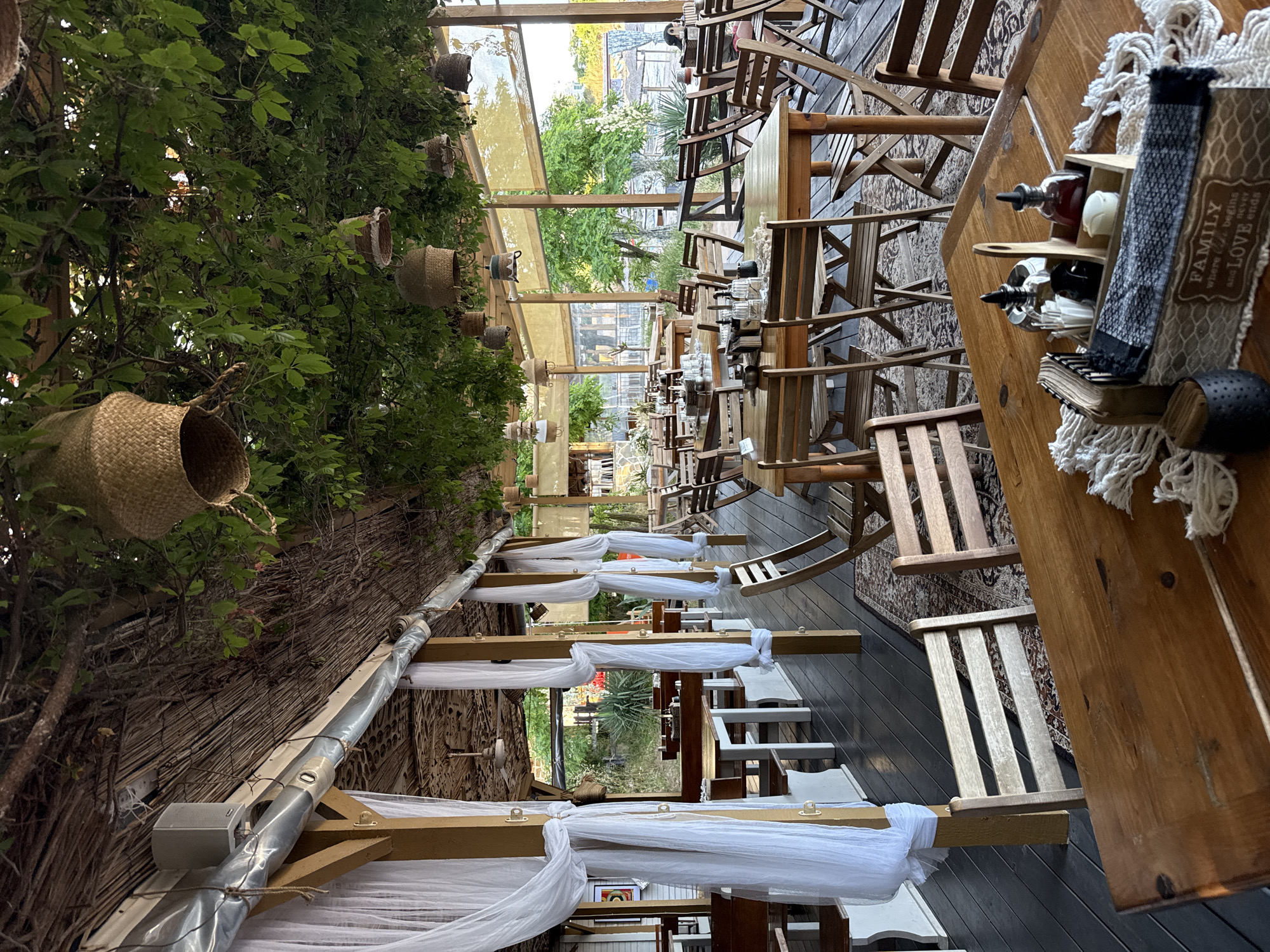 The image depicts an outdoor dining area at a restaurant located in Tsarevo, Bulgaria. The space is covered by a thatched roof and adorned with hanging greenery, creating a natural, shaded atmosphere. White sheer curtains are draped around wooden pillars, adding a touch of elegance. Numerous wooden tables and chairs are set up on a dark wooden deck, some with woven rugs underneath. In the foreground, a wooden table is set with a condiment caddy containing bottles, napkins, and sugar packets, alongside a decorative box with the inscription "FAMILY where life begins and LOVE never ends". The background shows more tables and chairs, glimpses of trees, and beyond that, a beach and the sea. The overall impression is that of a relaxed, rustic seaside eatery.