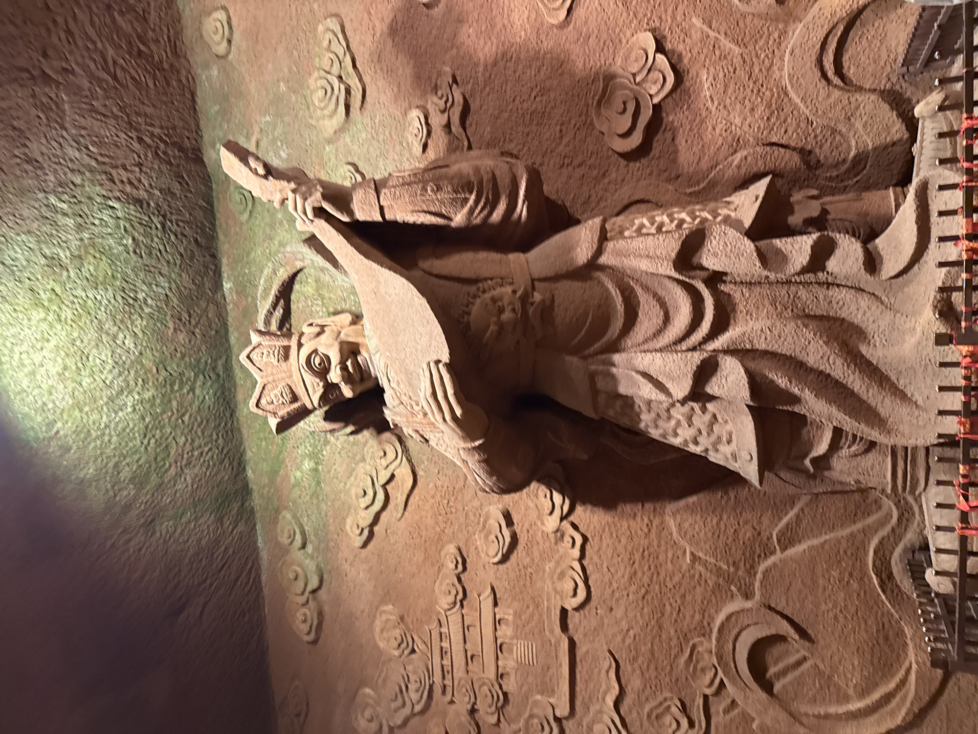 The image shows a stone carving in an unidentified location, likely a cave or temple. The carving depicts a figure with a crown, holding a flat object, possibly a tablet. The figure is surrounded by cloud and building motifs. The carving appears to be part of a larger work on a wall. The location is assumed to be in an area named Unknown in the city of Unknown.