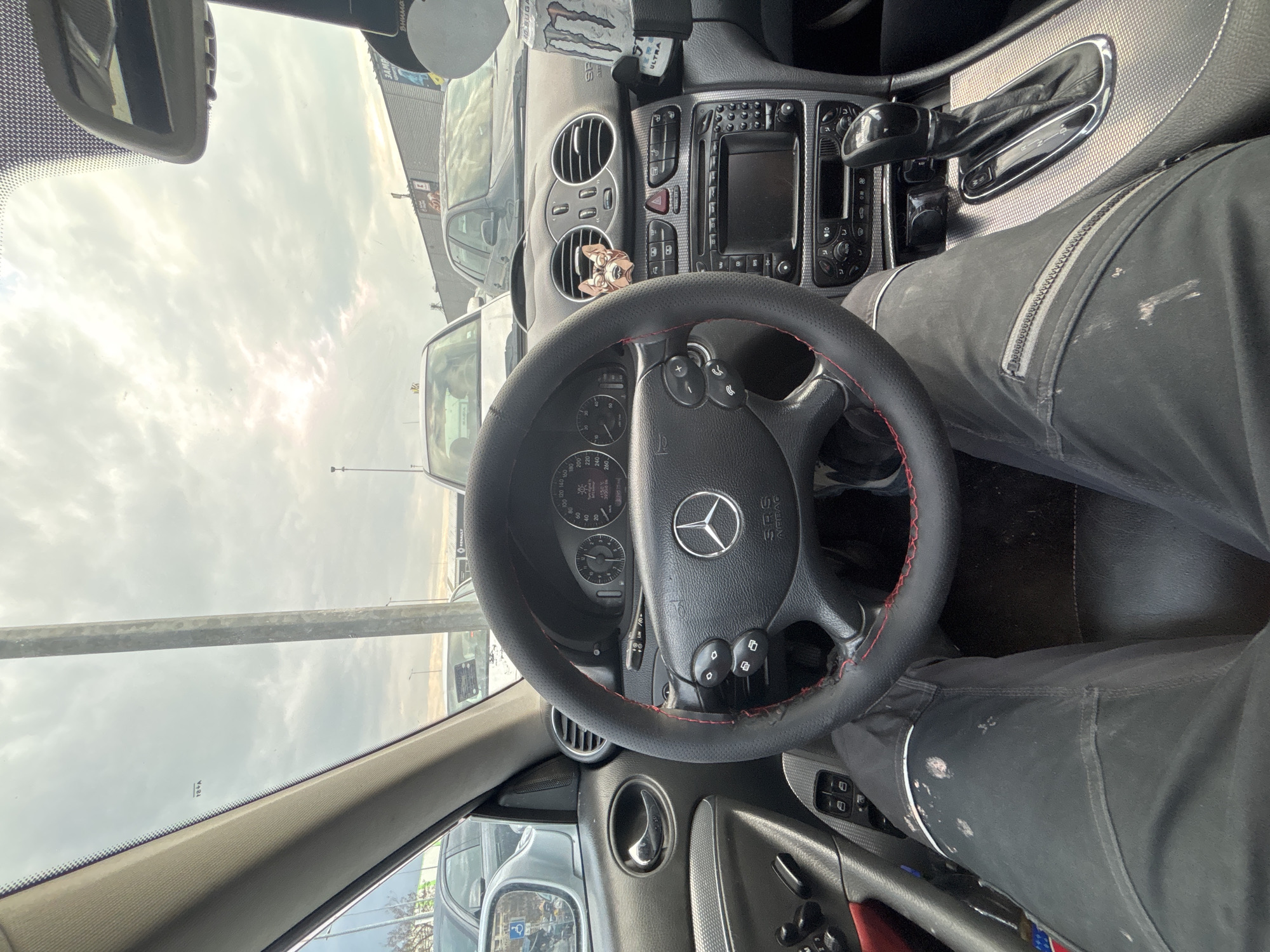 The image shows the interior of a Mercedes-Benz car, specifically the dashboard, steering wheel, and part of the driver's legs. The steering wheel has a red stitch detail. The car's dashboard is visible with the center console, vents, and infotainment system. The driver is wearing dark-colored trousers with some white stains. Outside the car, there are other parked vehicles and street elements, suggesting the location is an urban parking area in Burgas, Bulgaria. The sky is overcast. The photo appears to be taken from the driver's perspective while seated in the car.