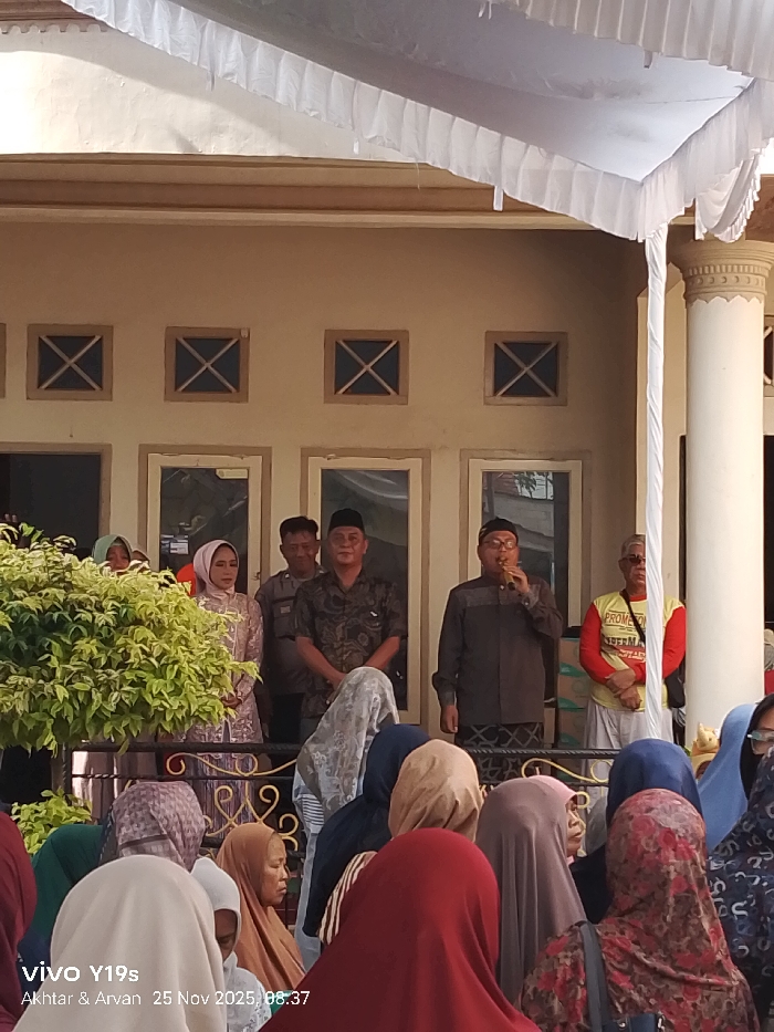 A gathering of people is shown in front of a two-story building. A woman in a hijab, a man in a black patterned shirt, and another man speaking into a microphone stand on the balcony. The building has windows with an X design in the top panels. A crowd of people, mostly wearing headscarves, stand below the balcony. A white canopy is strung above the balcony.