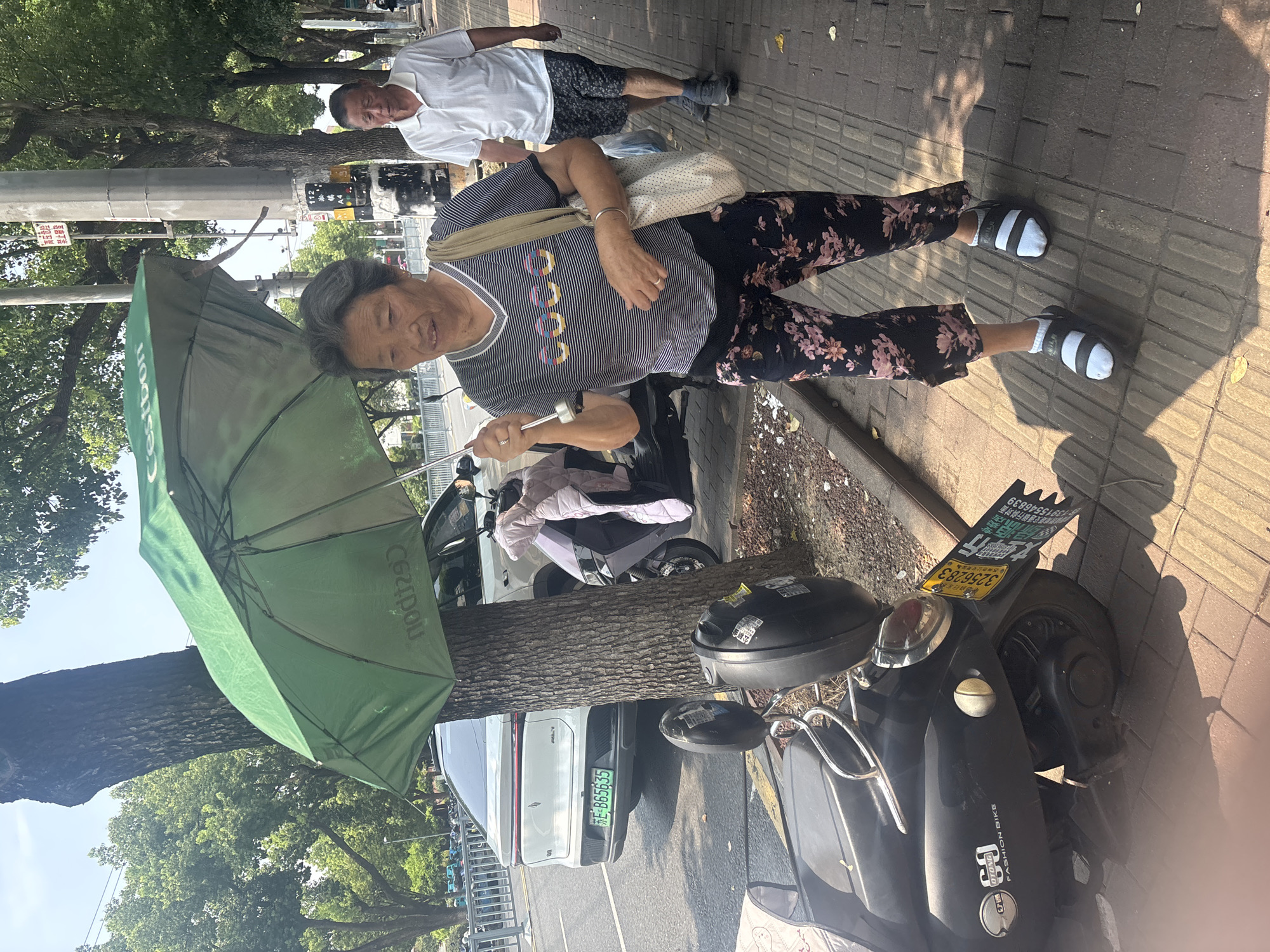 An elderly woman is walking on a sidewalk under a green umbrella, holding its handle with her right hand. She is wearing a striped t-shirt and floral pants. Beside her, a man in a white polo shirt and patterned shorts walks behind her. They are on a paved sidewalk lined with trees and a street with vehicles and parked scooters. The scene appears to be in an urban area with buildings and streetlights visible in the background. A white car with a green license plate and a black scooter are parked on the side of the street. The location is identified as Suzhou Industrial Park, China.