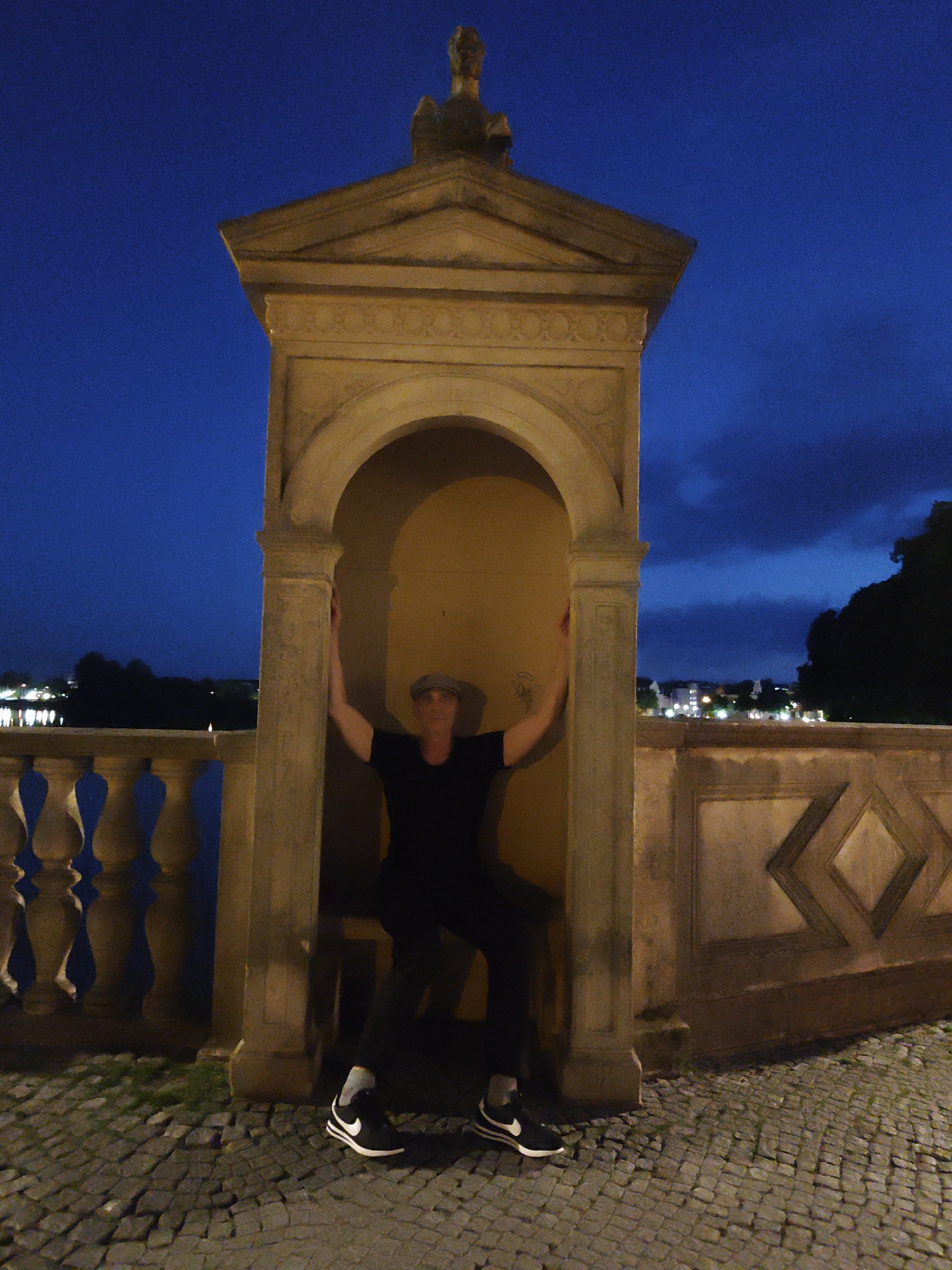 A person wearing a grey cap, a black t-shirt, black pants, and black Nike sneakers with white accents is posed inside a small, stone structure with arched openings. The structure appears to be part of a larger building or monument. The person is seated with their arms extended outwards, leaning against the interior walls of the structure. The scene is outdoors at dusk or night, with a deep blue sky visible. A stone balustrade with decorative spindles runs along a walkway in the foreground, and a cobblestone path is visible on the ground. In the background, lights from a city or town can be seen across a body of water, with trees silhouetted against the sky. This scene is located on the Schwerin Castle Island (Schlossinsel) in Schwerin, Germany, specifically on the terrace overlooking the Old Garden (Alter Garten).